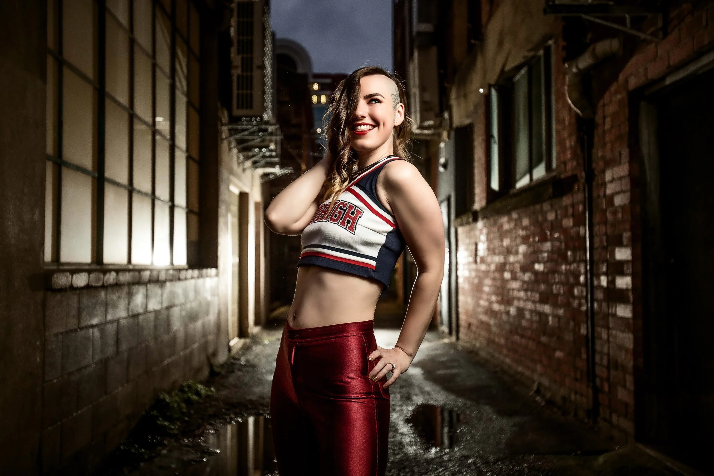 A woman with wavy hair, a tattoo on her head, and bright red lipstick, standing in an urban alleyway at night. She is wearing a cheerleader-style top and shiny red pants, smiling and touching her hair with one hand.