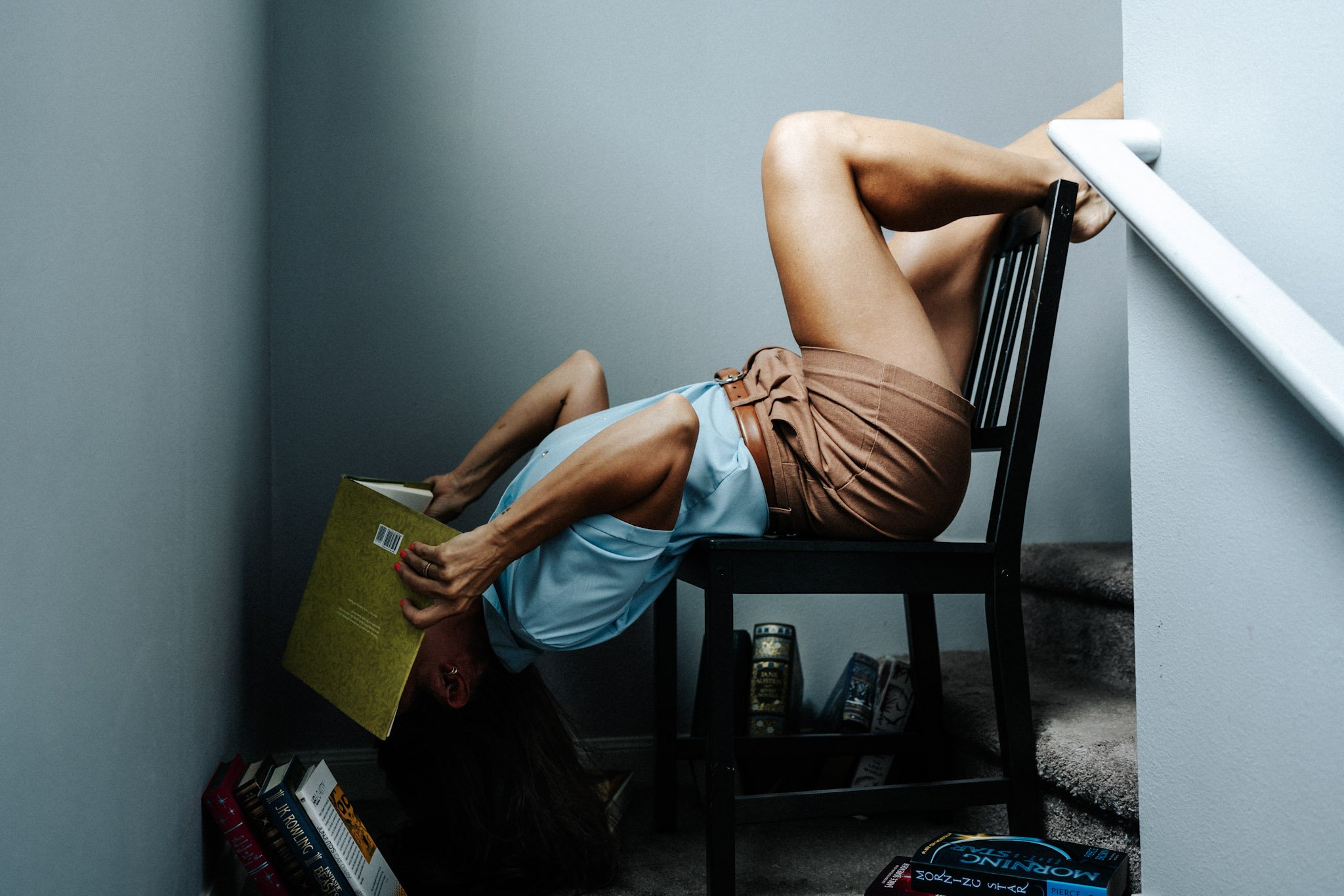 A woman with long hair is sitting upside down on a black chair with her head resting on the floor and holding a yellow book. She is wearing a light blue top and brown shorts. The scene appears to be in a room with gray walls and a gray carpeted floor, with various books stacked on the floor and a small bookshelf.