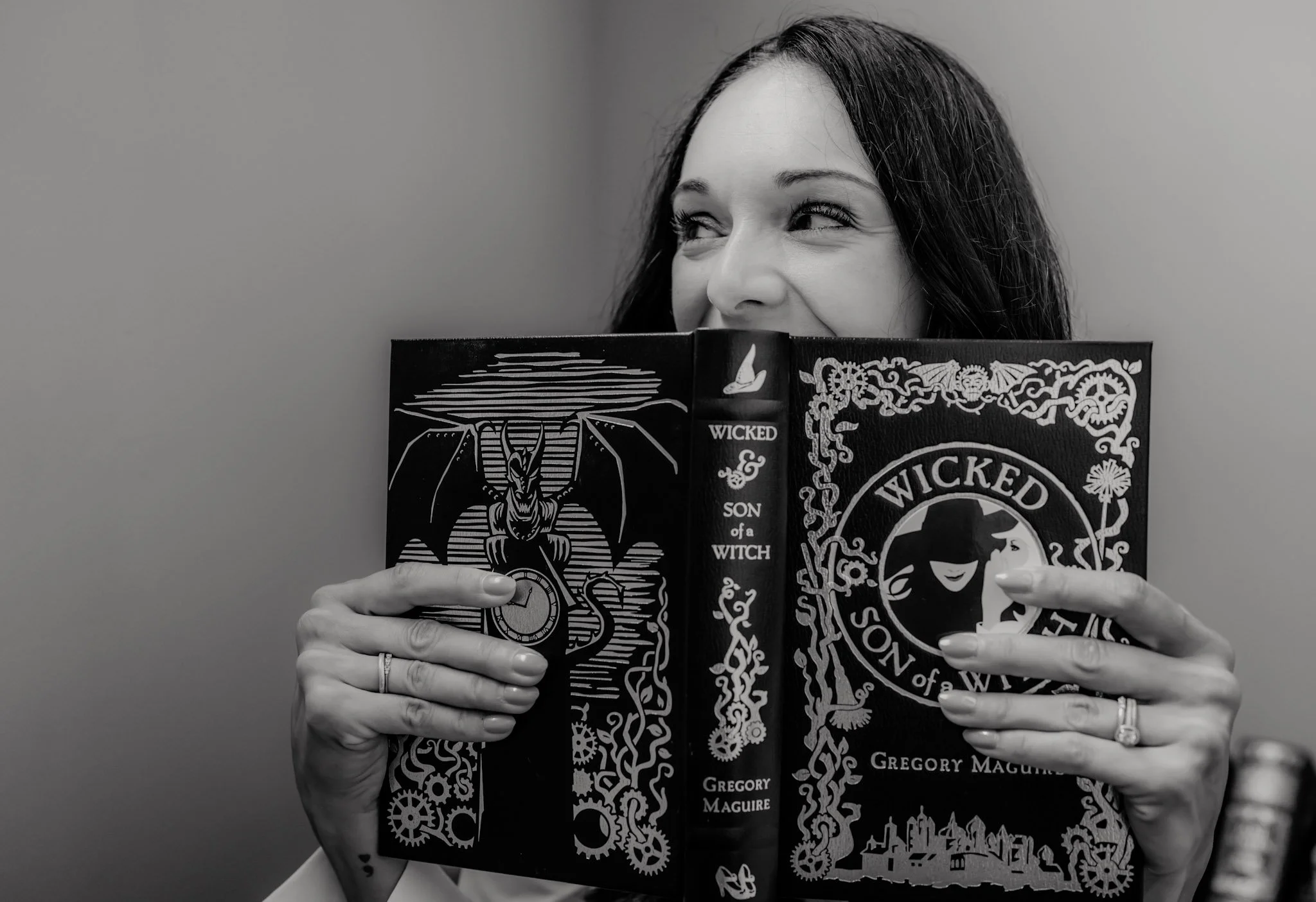 A woman with dark hair holding a book titled "Wicked" by Gregory Maguire, partially covering her face, with her eyes looking to the side, against a plain background.