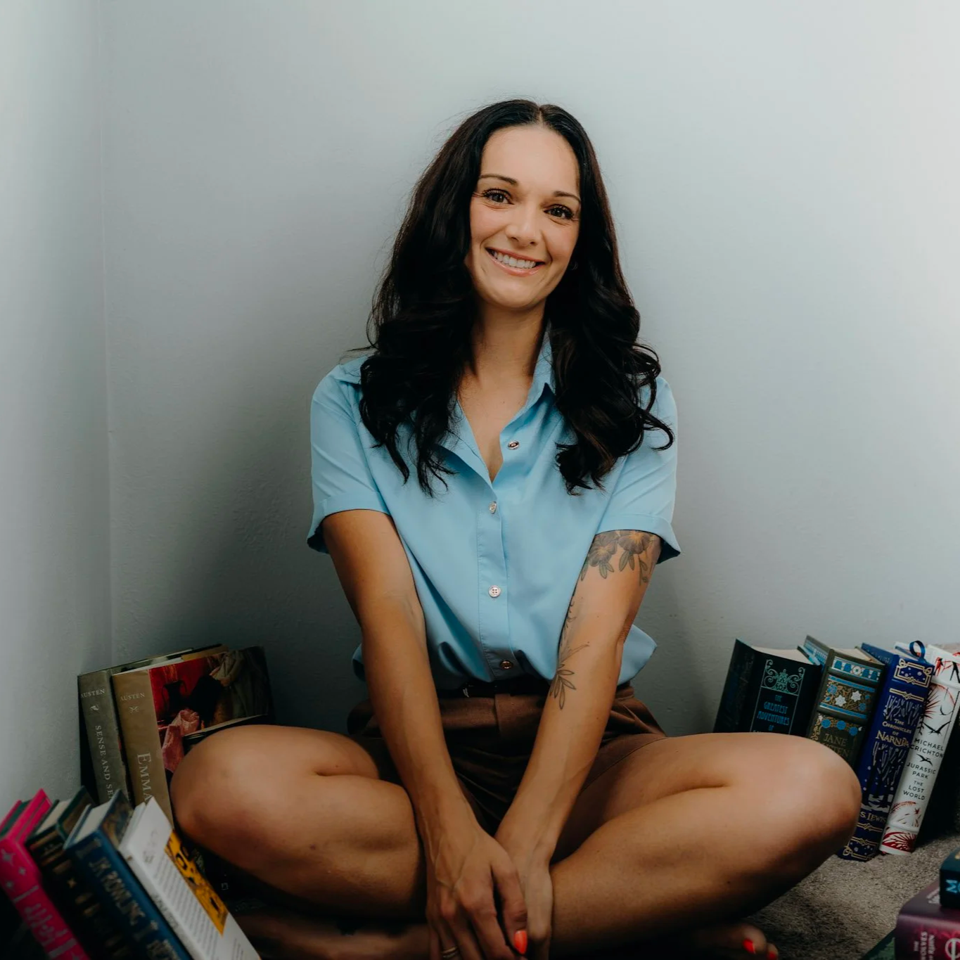 A woman sitting cross-legged on the floor surrounded by books, smiling at the camera, in a room with plain white walls.