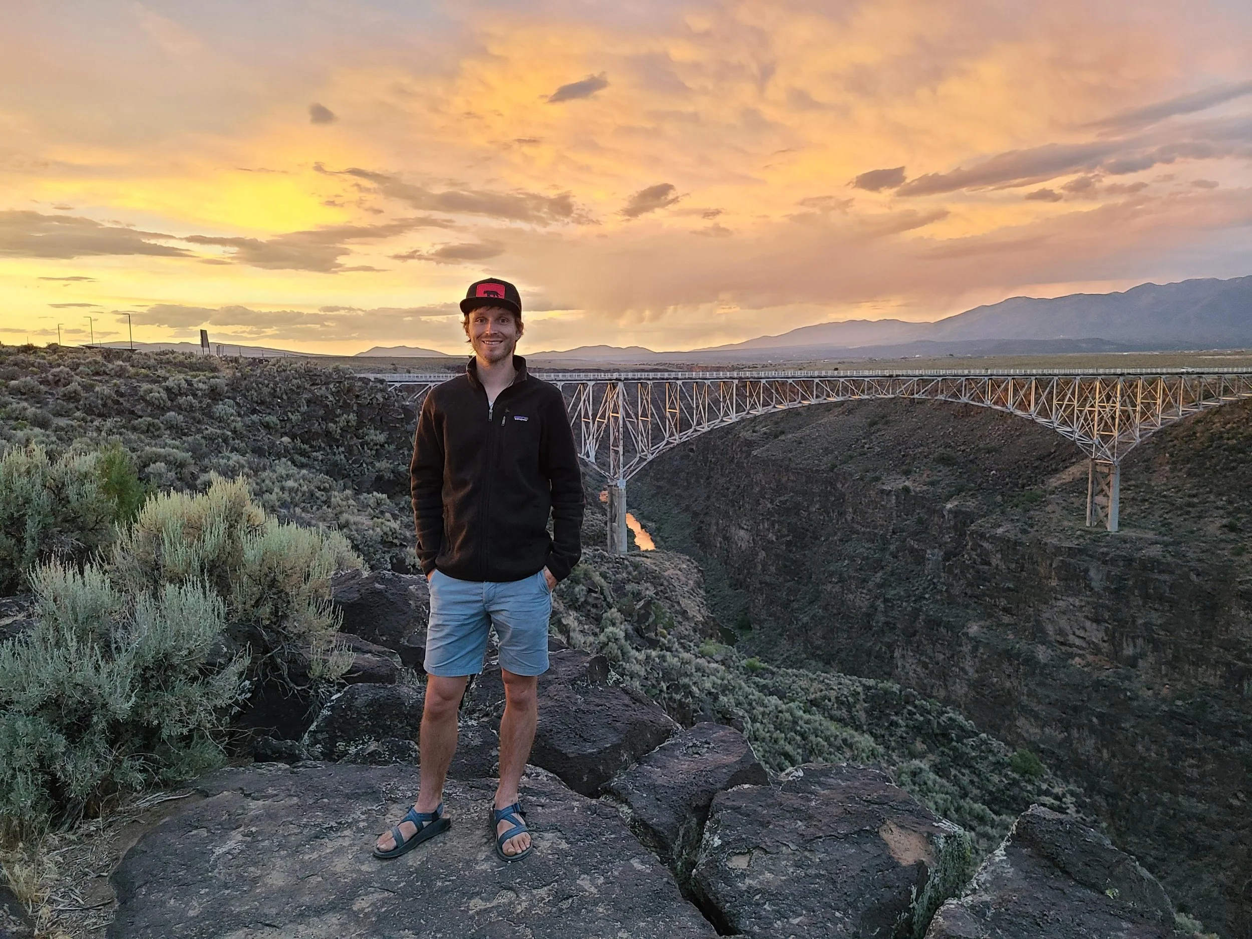A man standing on rocks near a canyon with a bridge and mountains in the background during sunset.