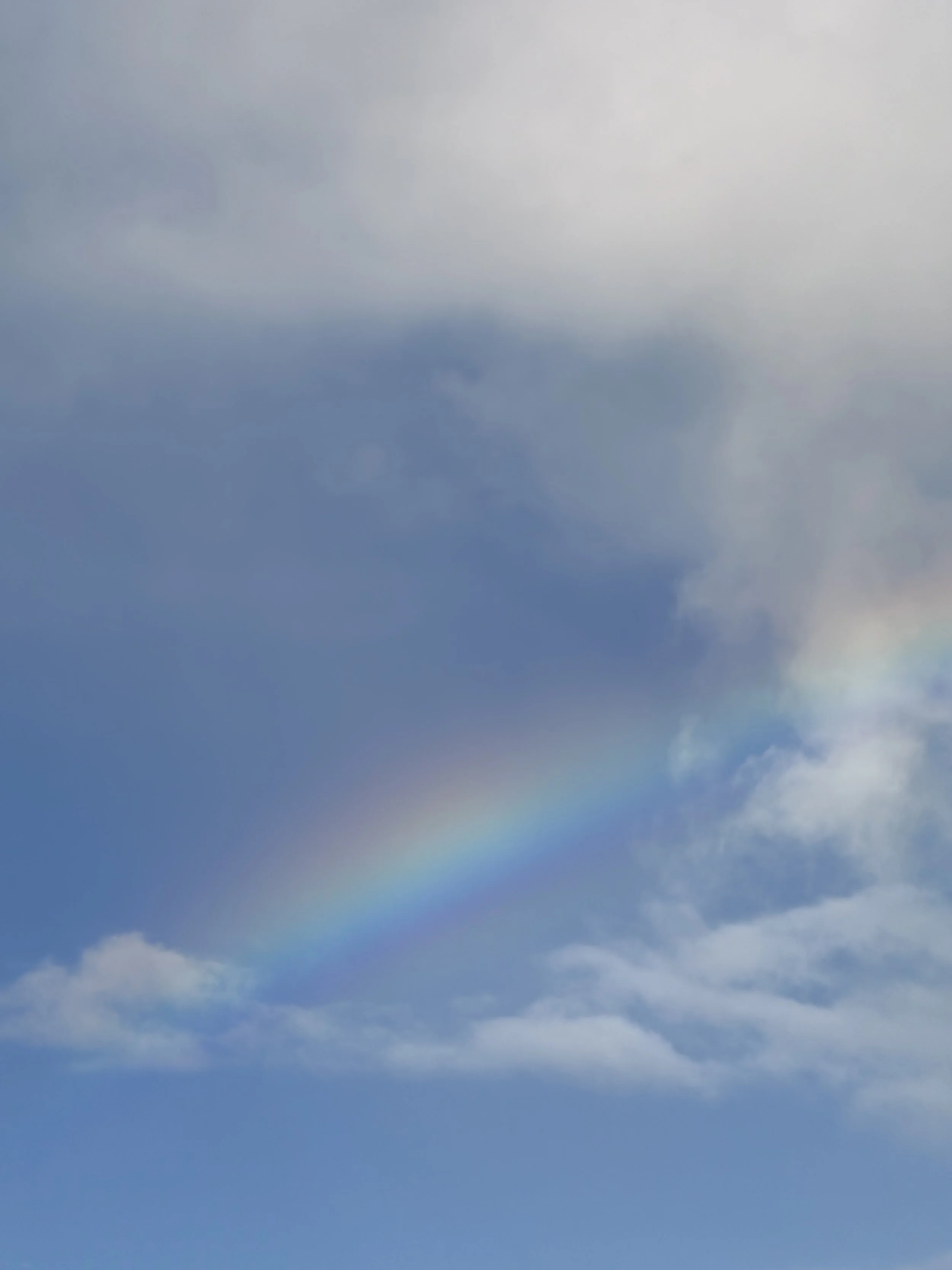 Cloudy blue sky with a faint rainbow and some wispy clouds.