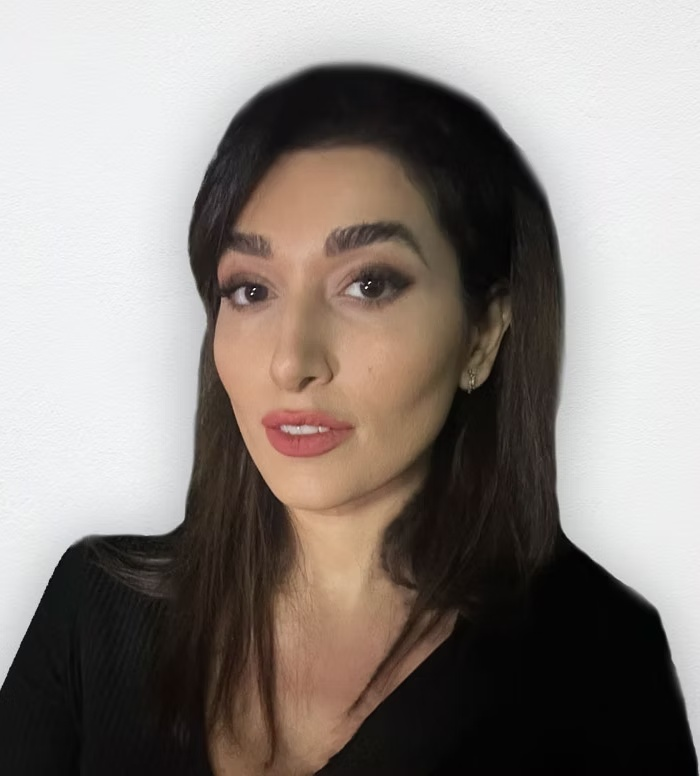 A woman with long dark hair, wearing makeup and earrings, looking into the camera against a plain white background.