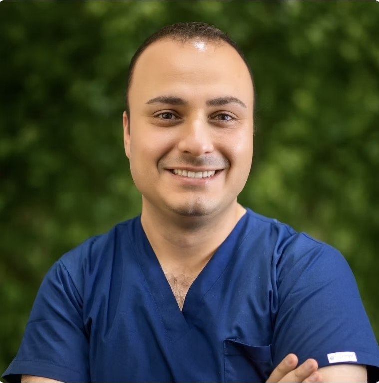 A smiling man with short dark hair, wearing blue scrubs, standing outdoors with a blurred green background.