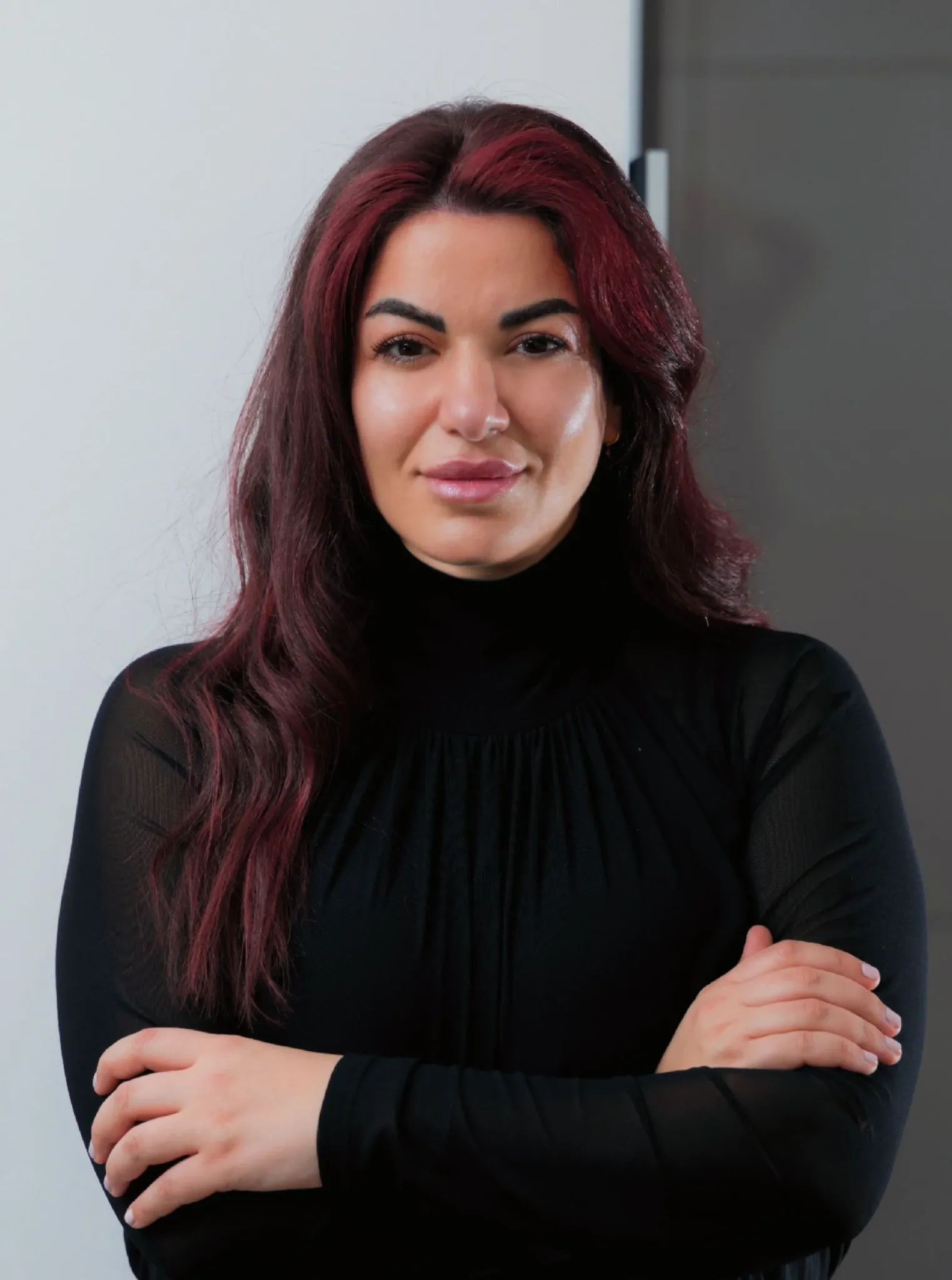 Close-up of a woman with dark and red hair, wearing a black top, with arms crossed, standing against a plain light and dark background.