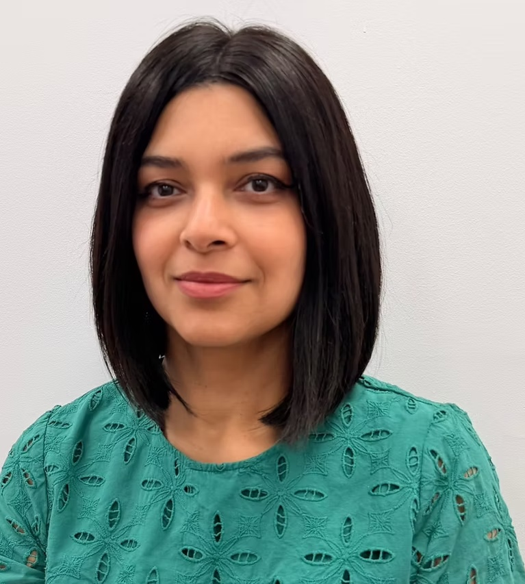 A young woman with shoulder-length black hair, wearing a green top with a pattern of oval-shaped cutouts, standing against a plain white wall.
