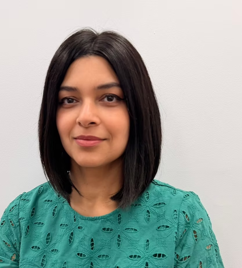 A woman with shoulder-length black hair and a slight smile, wearing a green blouse with eyelet embroidery, standing against a plain white wall.