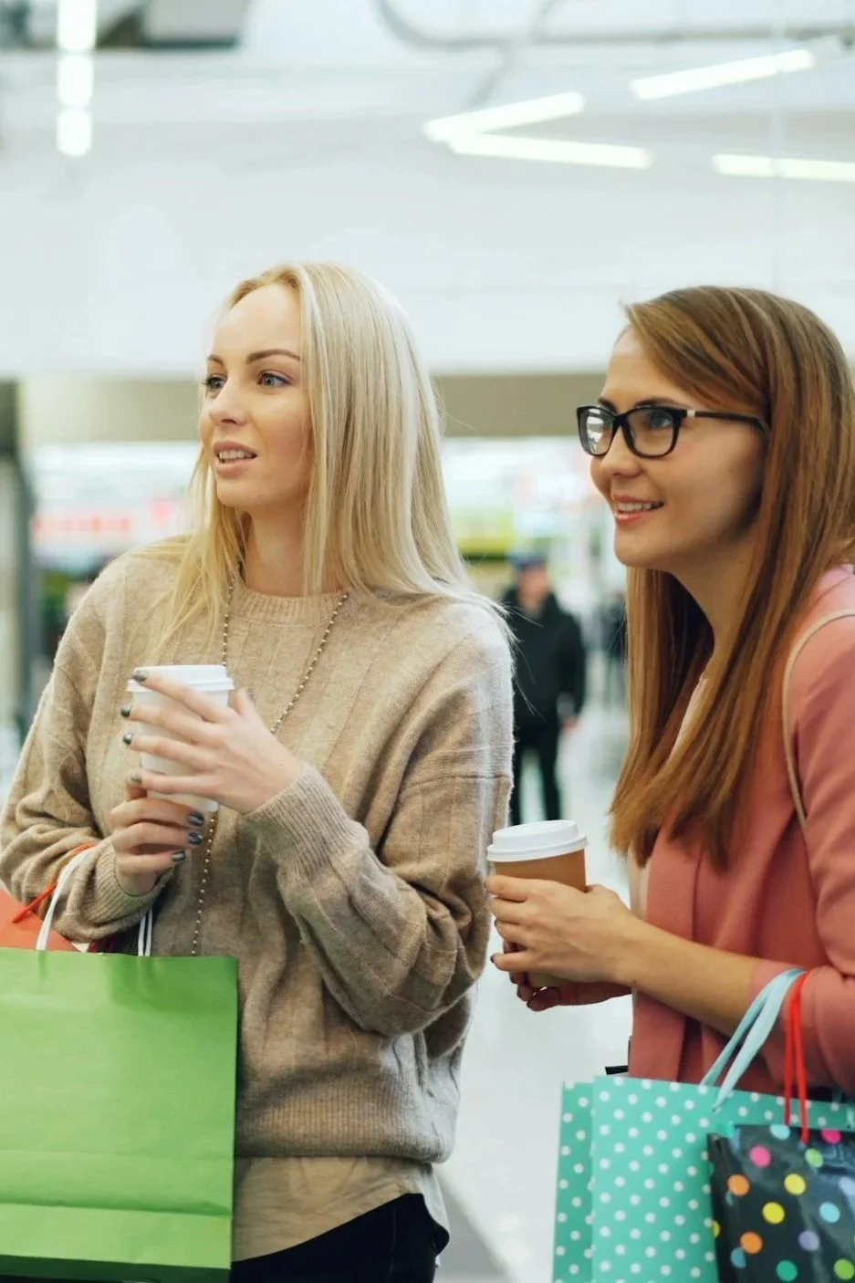 Two women holding coffee cups and shopping bags in a shopping mall.