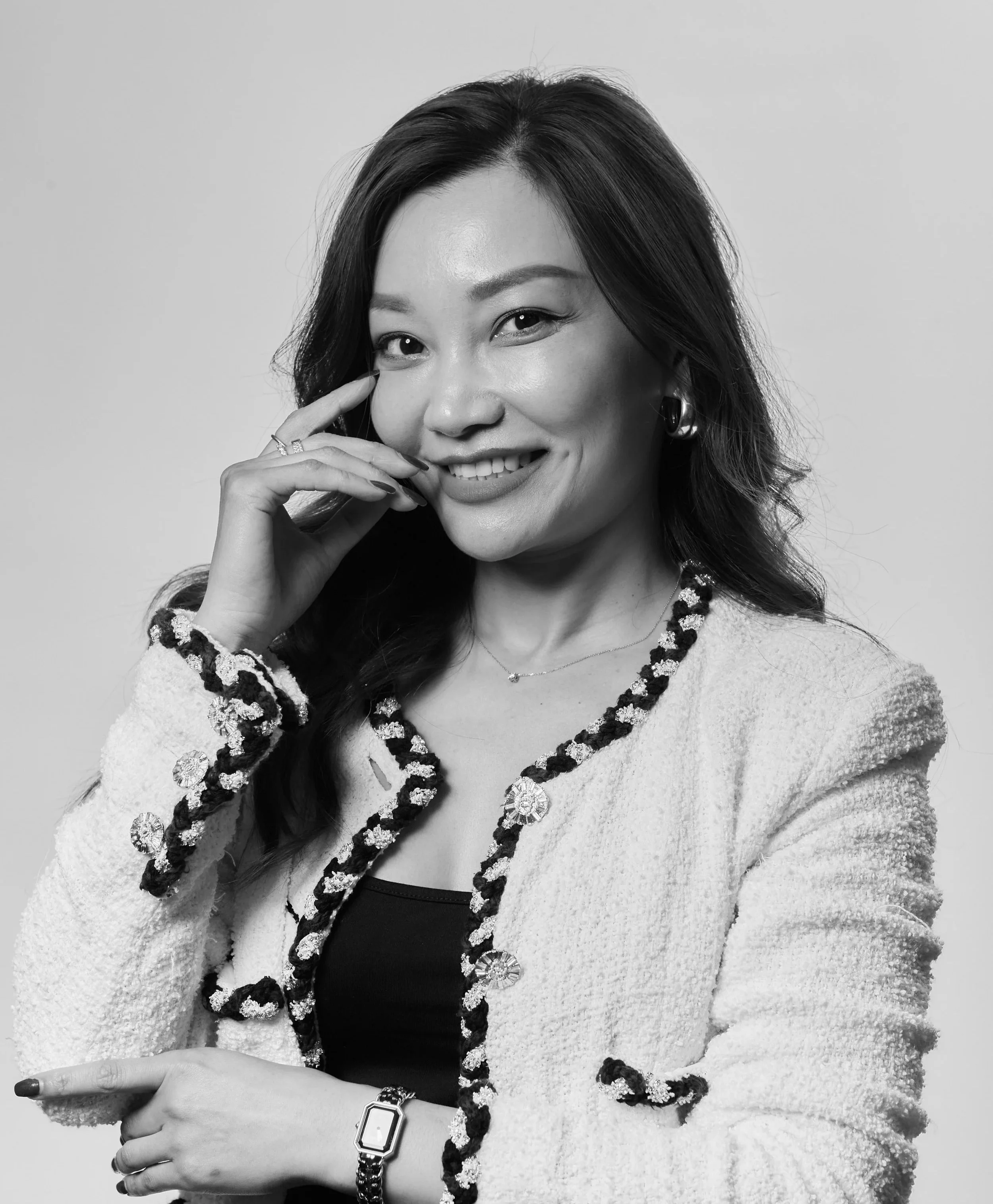 Black and white photograph of a smiling woman with dark hair, wearing a watch, rings, and a tailored blazer with decorative trim, posing with one hand near her face and looking at the camera.