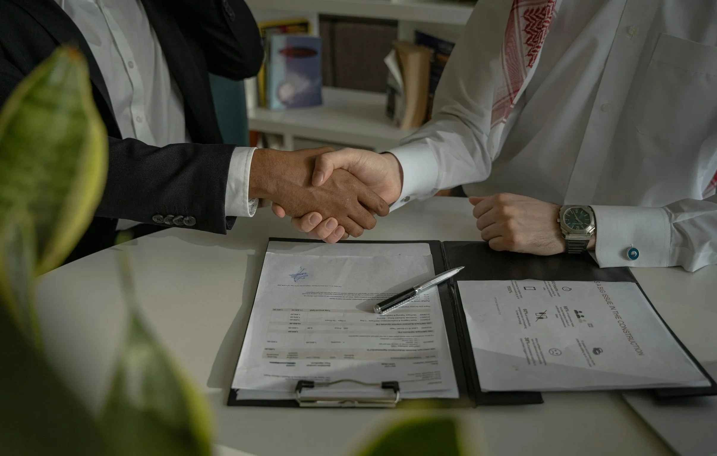Close-up of a handshake between two people in formal attire, possibly during a meeting or signing agreement, with documents, a pen, and office supplies on the table.