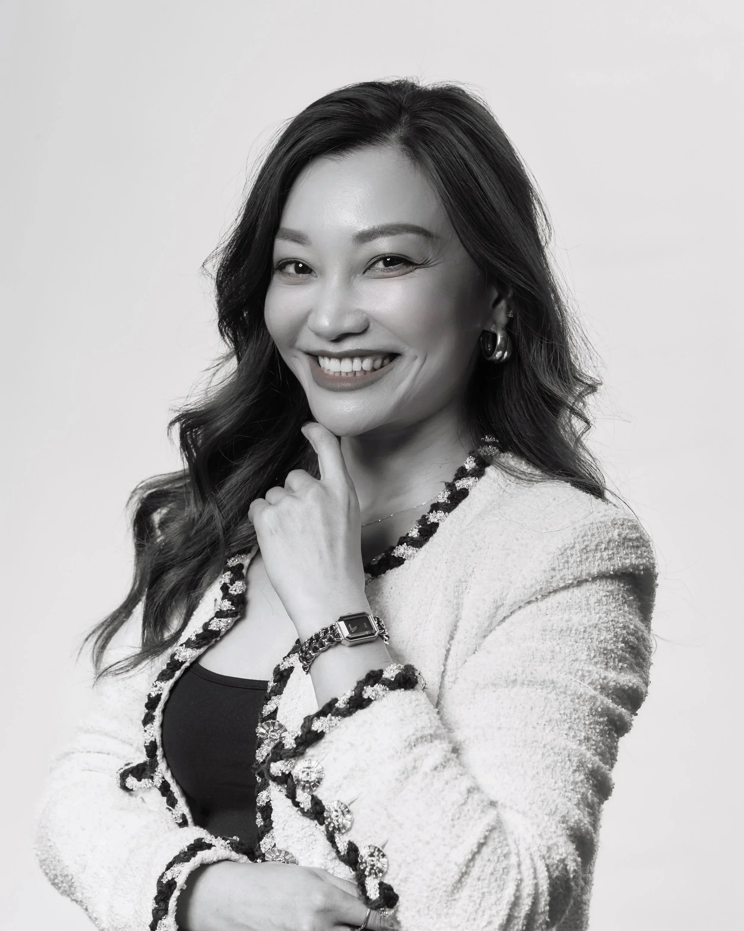 Smiling woman with long wavy hair, wearing a textured cardigan with decorative buttons, a black top, a watch, and earrings, posing in front of a plain background.