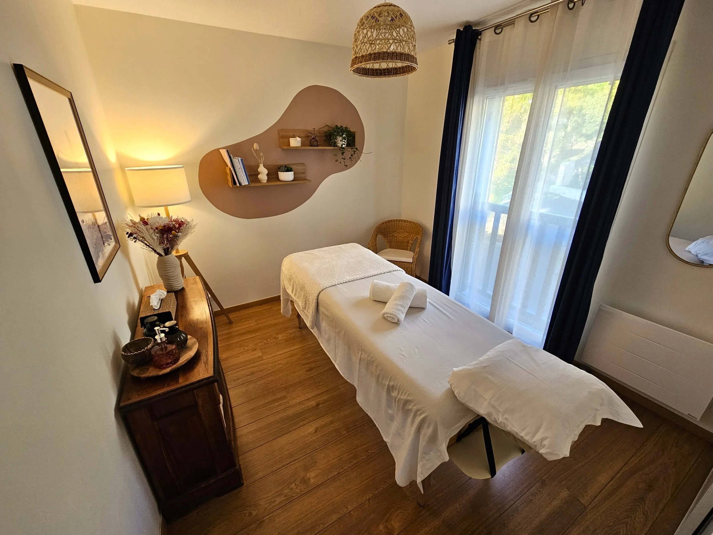 salle de soin, table de massage avec un drap blanc et un coussin blanc pour la tête. Des serviettes en coton, un fauteuil en rotin dans un coin de pièce, des rideaux bleus marines