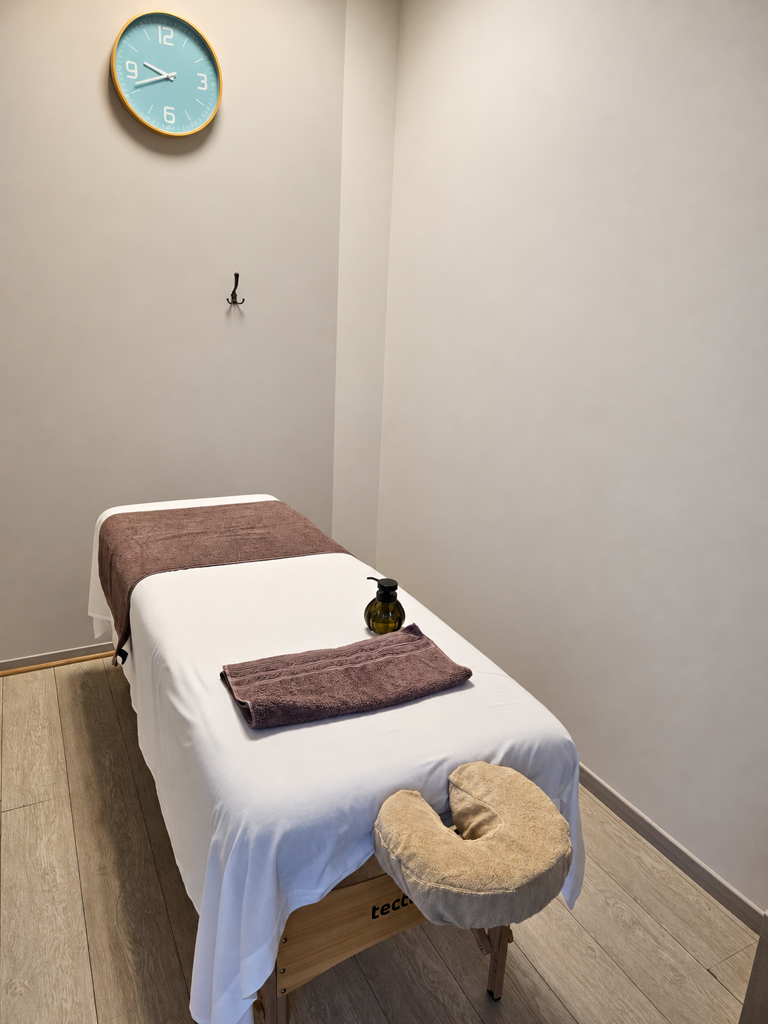 Cabine de massage avec table recouverte d'une serviette blanche, tapis de massage marron, bouteille d'huile, nappe marron, mur gris clair, horloge bleue, crochet noir au mur, coussin de massage beige sur la tête de la table.
