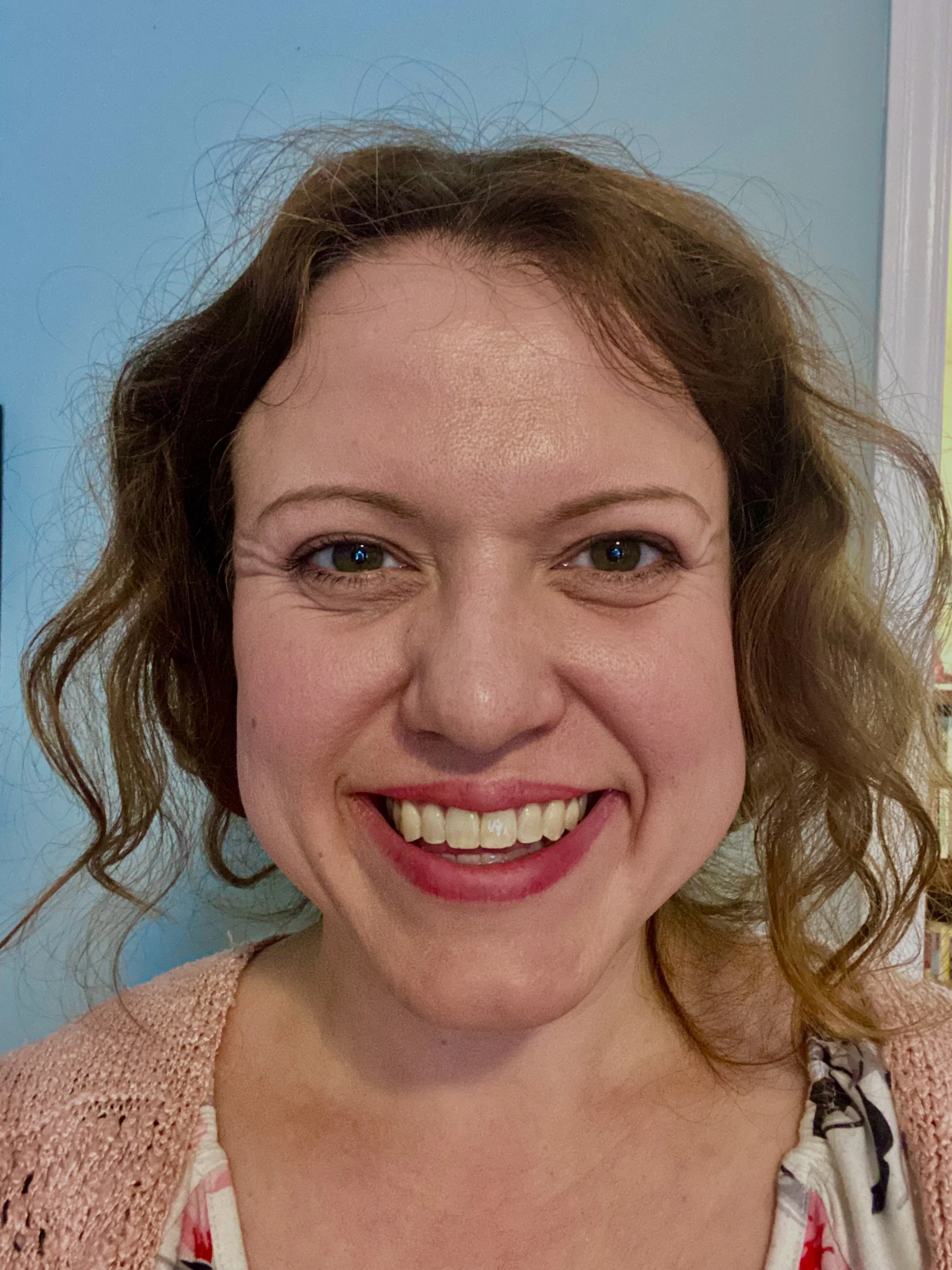 A smiling woman with curly brown hair and green eyes, wearing a light pink cardigan and a patterned shirt, standing in front of a blue wall.