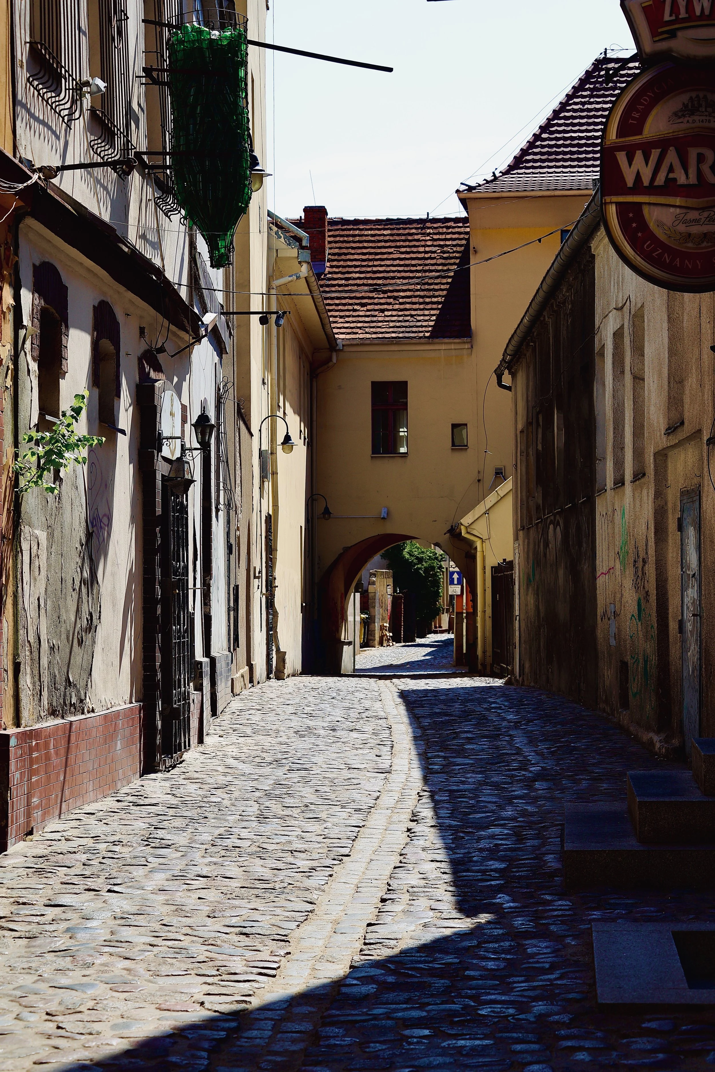 A narrow cobblestone street in an old European town, with old building walls on both sides and the street leading under an archway into brighter sunlight.