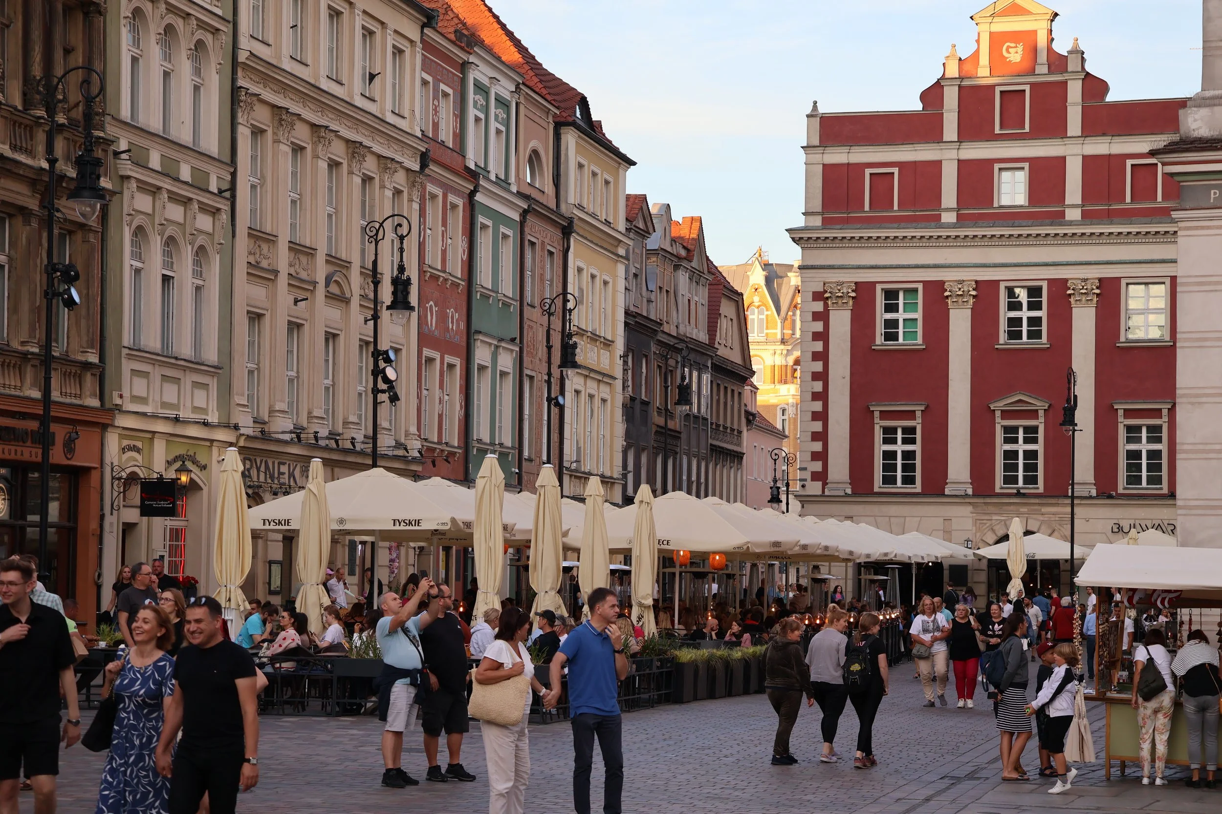 Crowd of people in a lively outdoor city square with historic buildings and outdoor cafes with umbrellas.