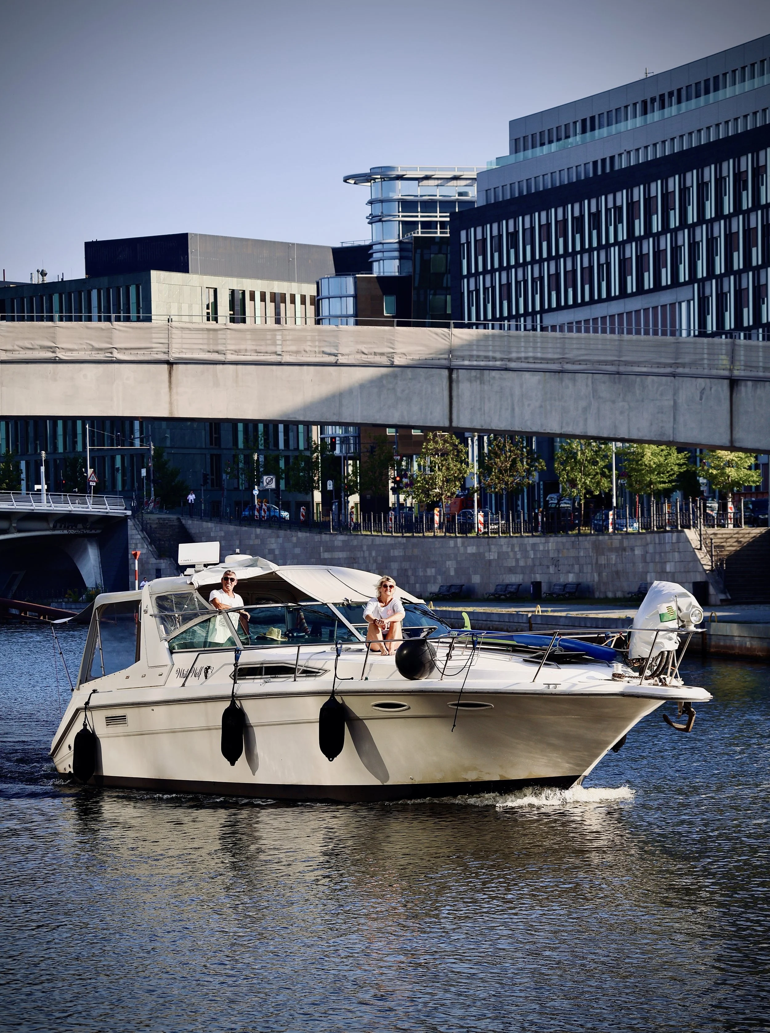 A white yacht with two people on deck, sailing on a river or canal with modern buildings and a pedestrian bridge in the background.