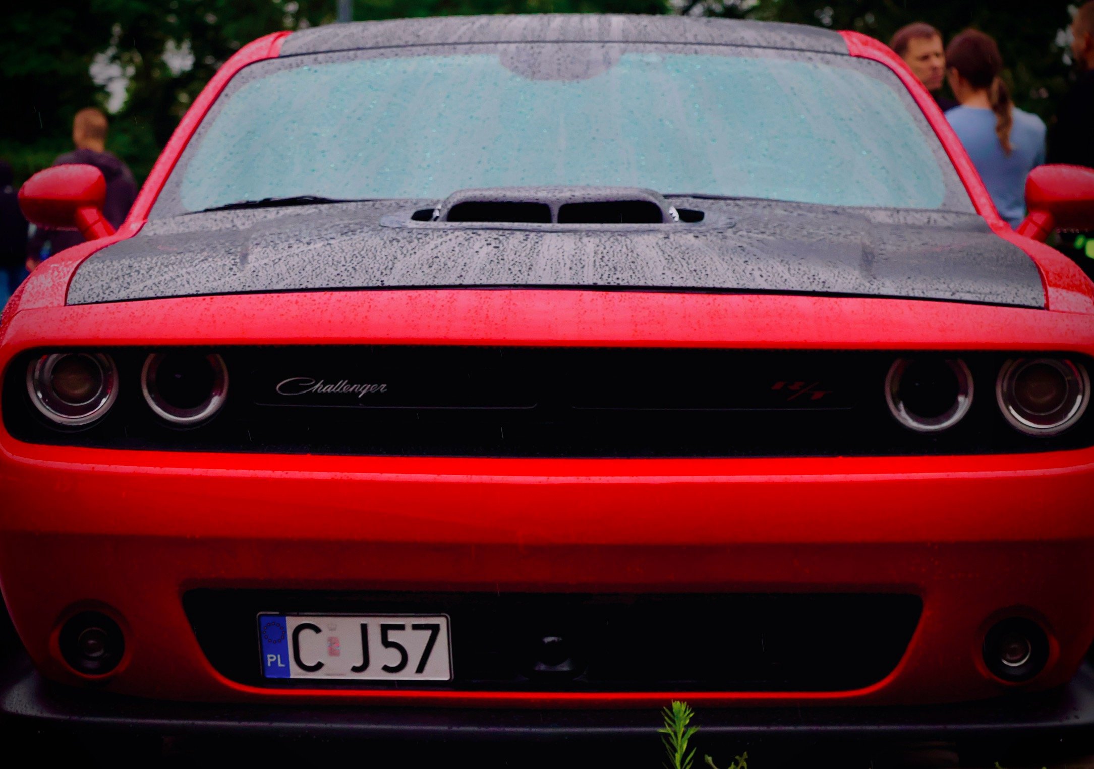 Front view of a red Dodge Challenger car with a black hood scoop, rainy weather, and a European license plate, with two women and some trees in the background.