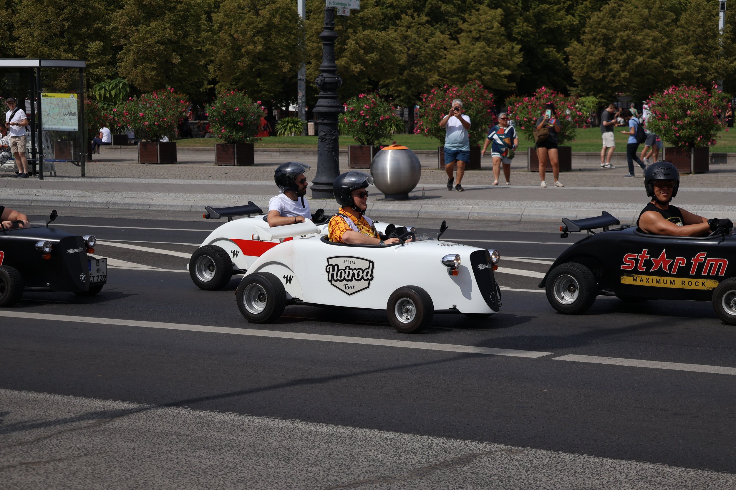 People riding small, white, hot rod-style cars on a city street, with pedestrians walking and taking photos, and trees and flowers lining the sidewalk in the background.