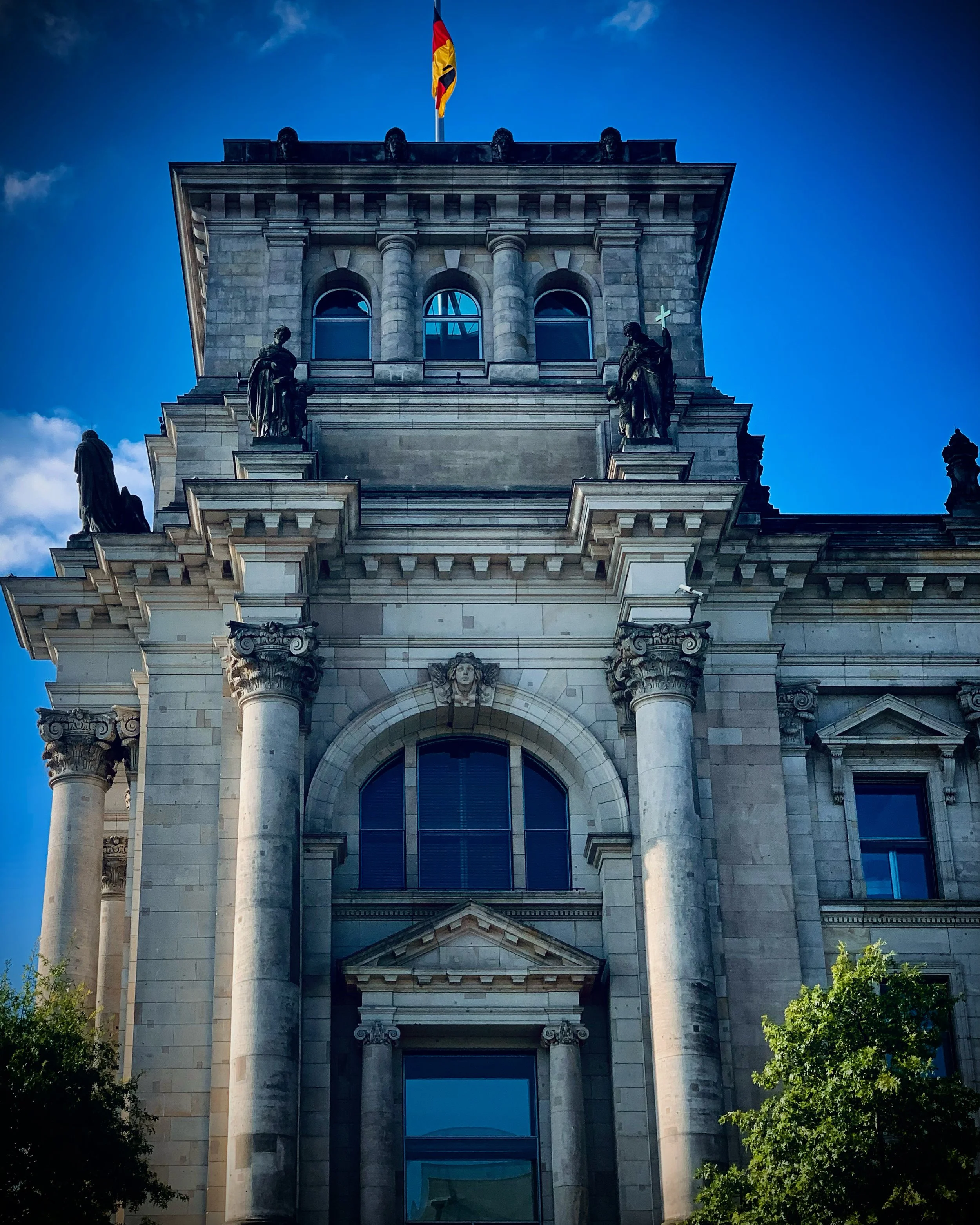 Front view of a historic building with classical architecture, including columns, statues, and a flag on top, under a clear blue sky.