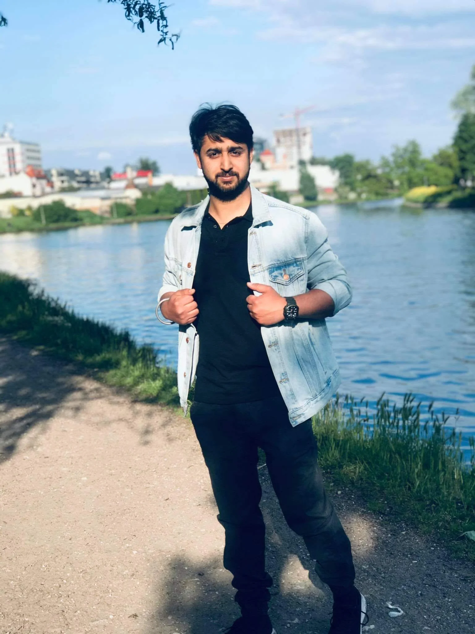 A young man with dark hair and a beard standing outdoors near a body of water with buildings and trees in the background, wearing a denim jacket over a black shirt, black pants, and sneakers, with his hands holding the jacket's lapels.