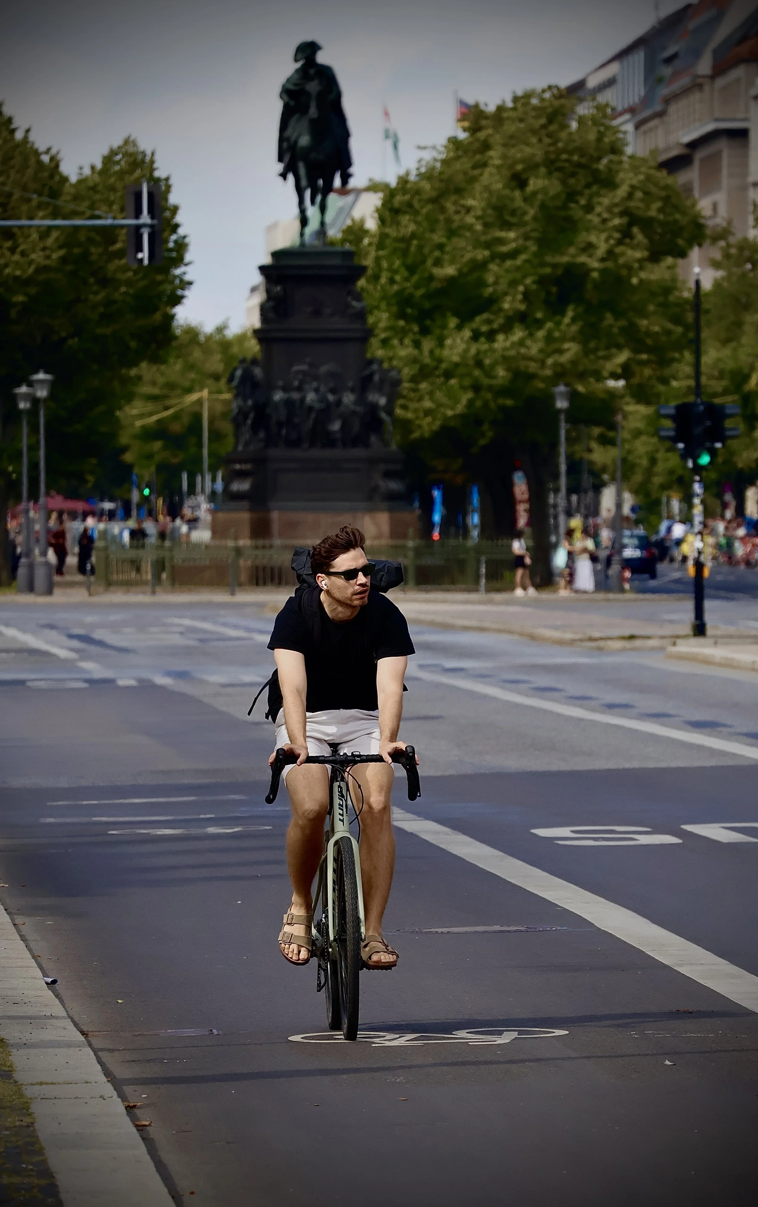 A man riding a bicycle on a city street during the daytime, with a statue and buildings in the background.