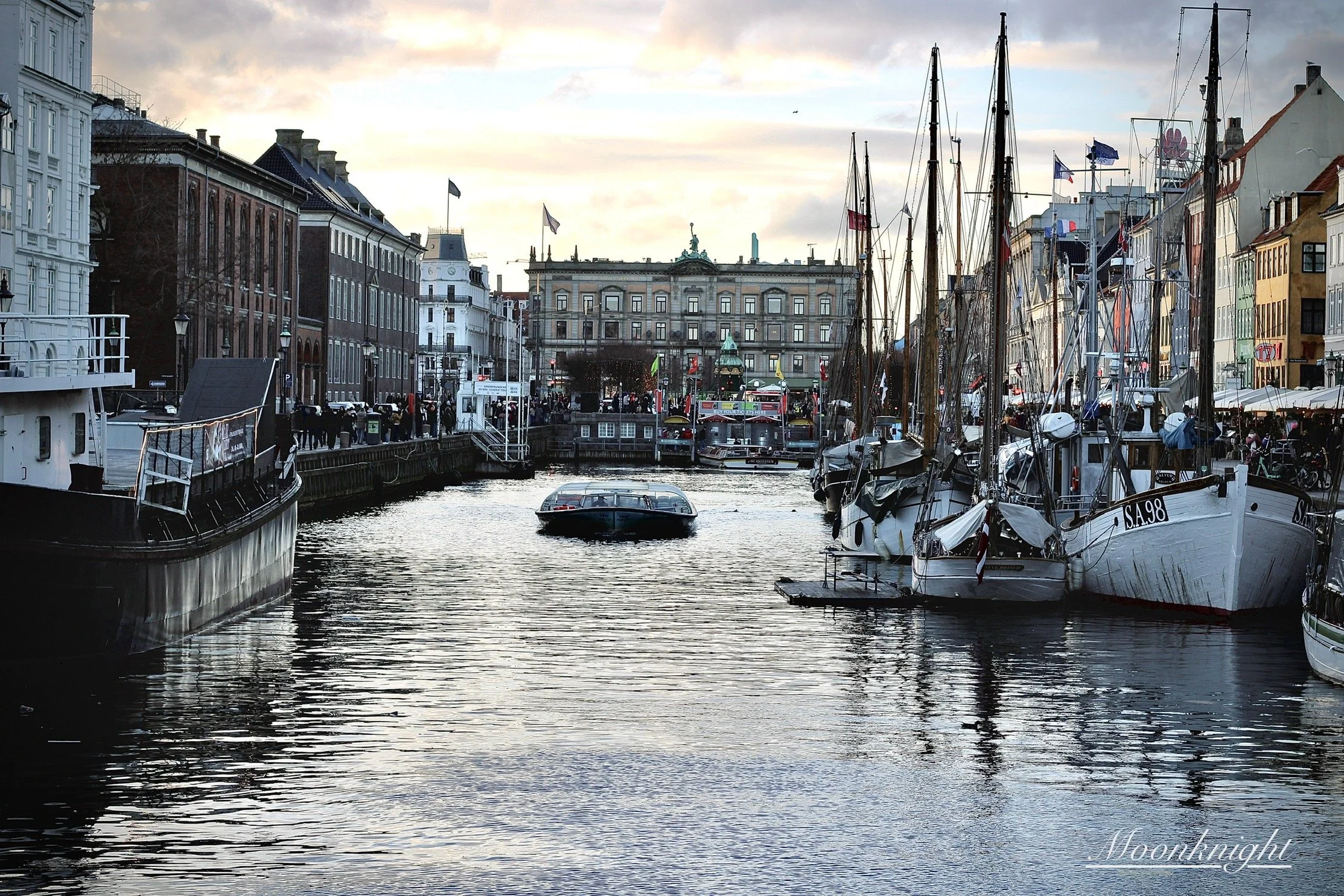 A harbor scene with sailboats docked along a canal, historic buildings on either side, and a large building with flags in the background, under a cloudy sky during sunset.