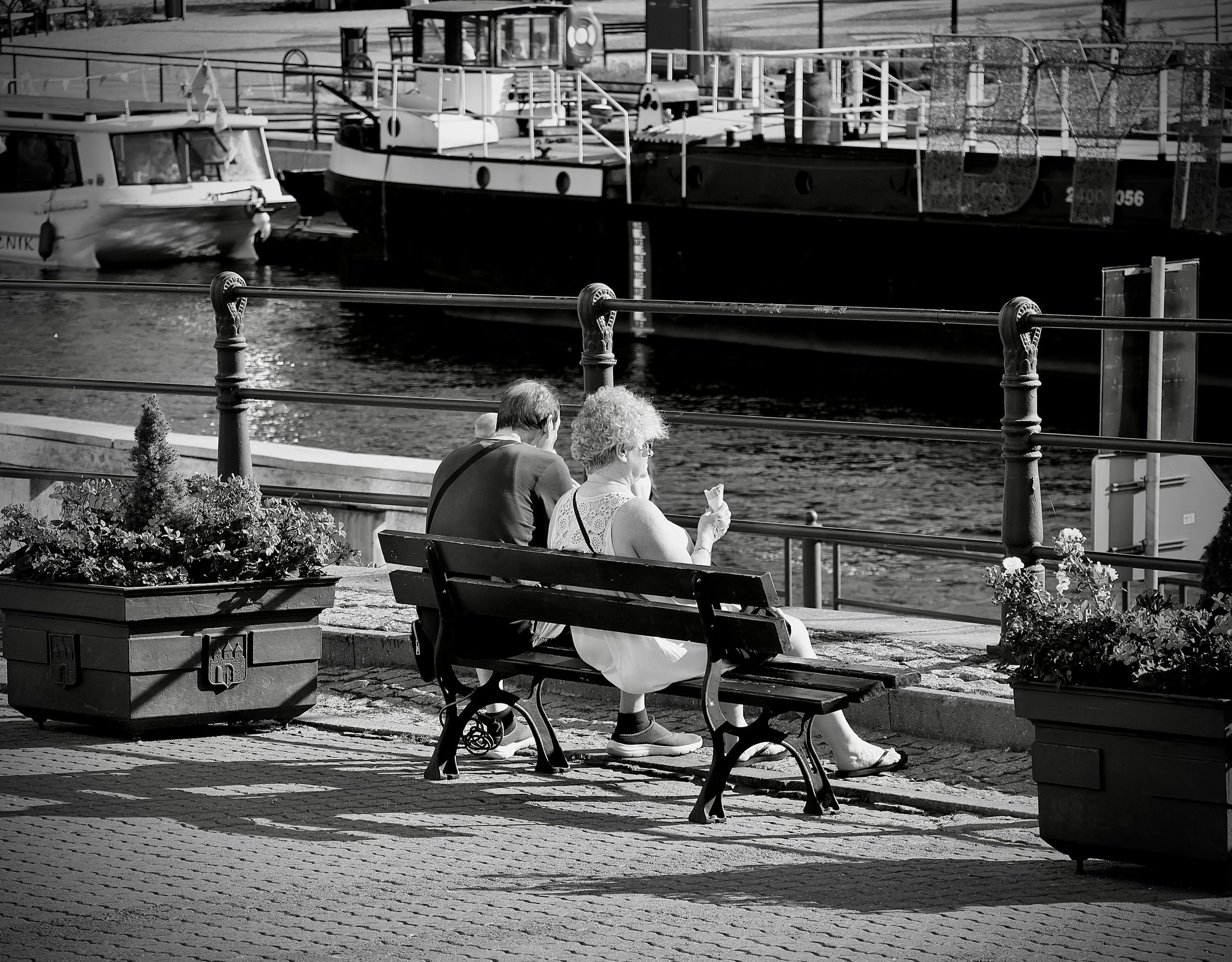 Two elderly women sit on a park bench near water, with boats visible in the background. They are facing sideways, one with curly hair holding a paper or menu, and the other with short hair.