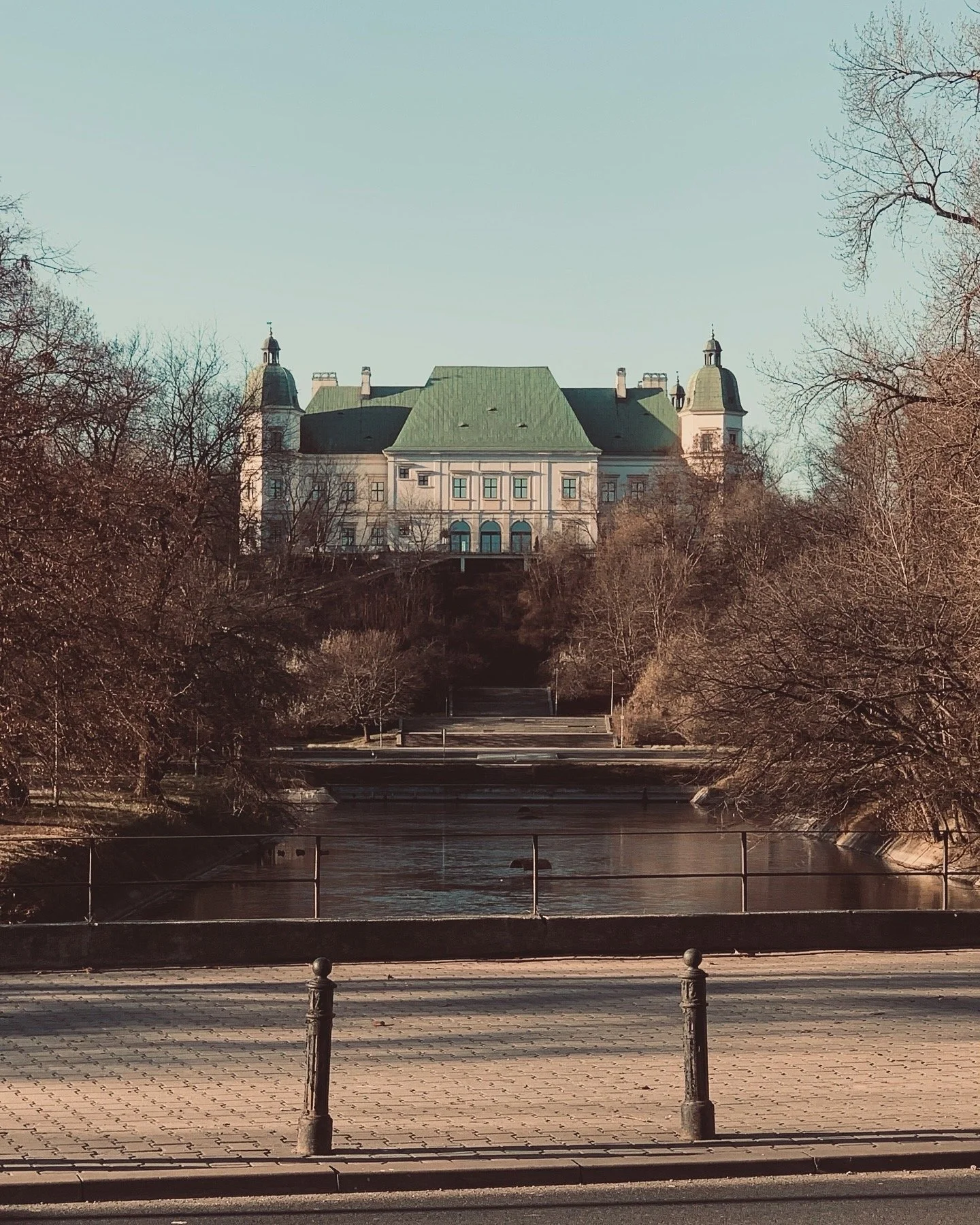 A large historic building with a green roof, situated on a hill, surrounded by leafless trees, with a river and a paved promenade with metal posts in the foreground.