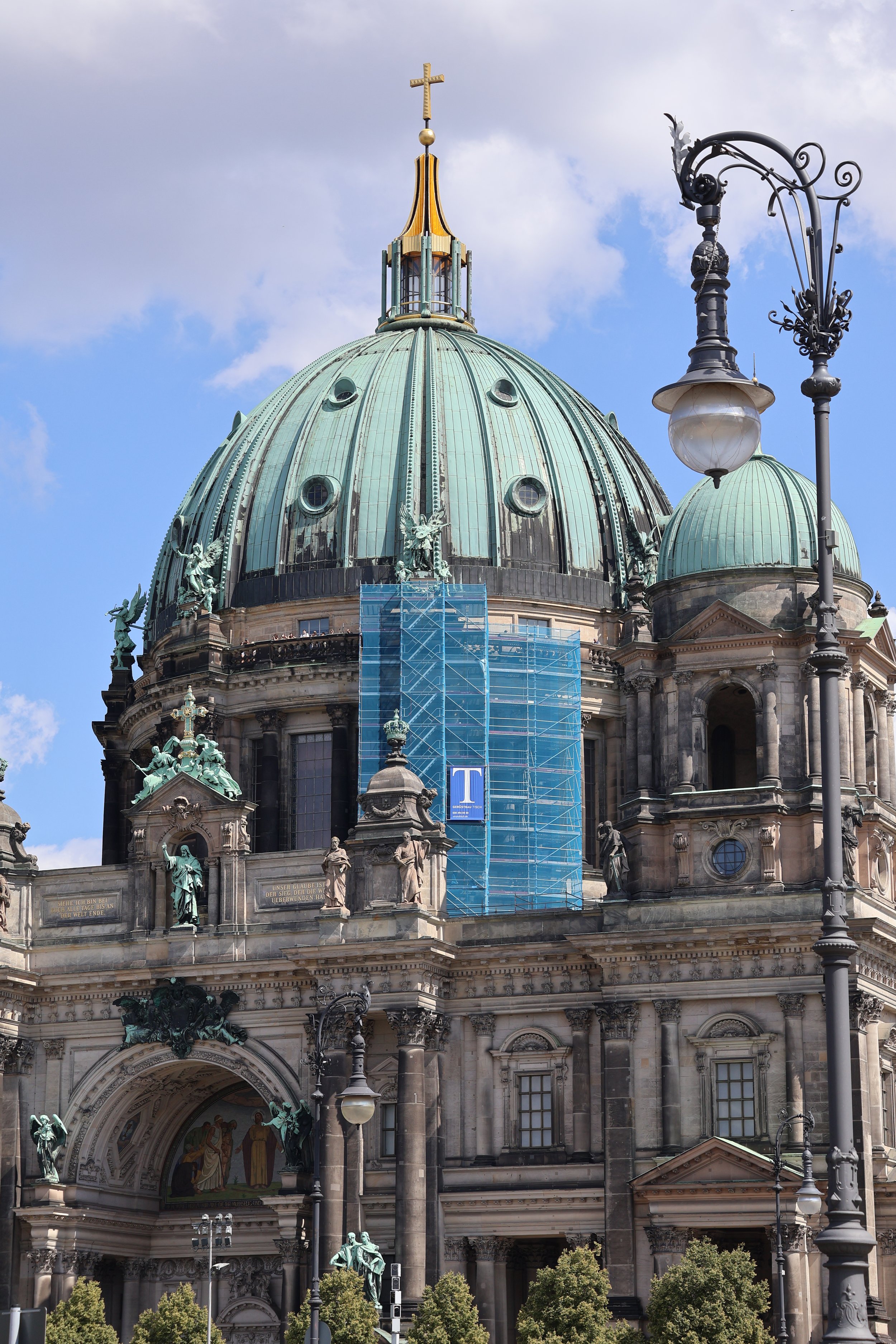 A historic building with a large dome and smaller dome, adorned with statues and a cross at the top, under a partly cloudy sky. There is a street lamp in the foreground and green trees at the bottom.