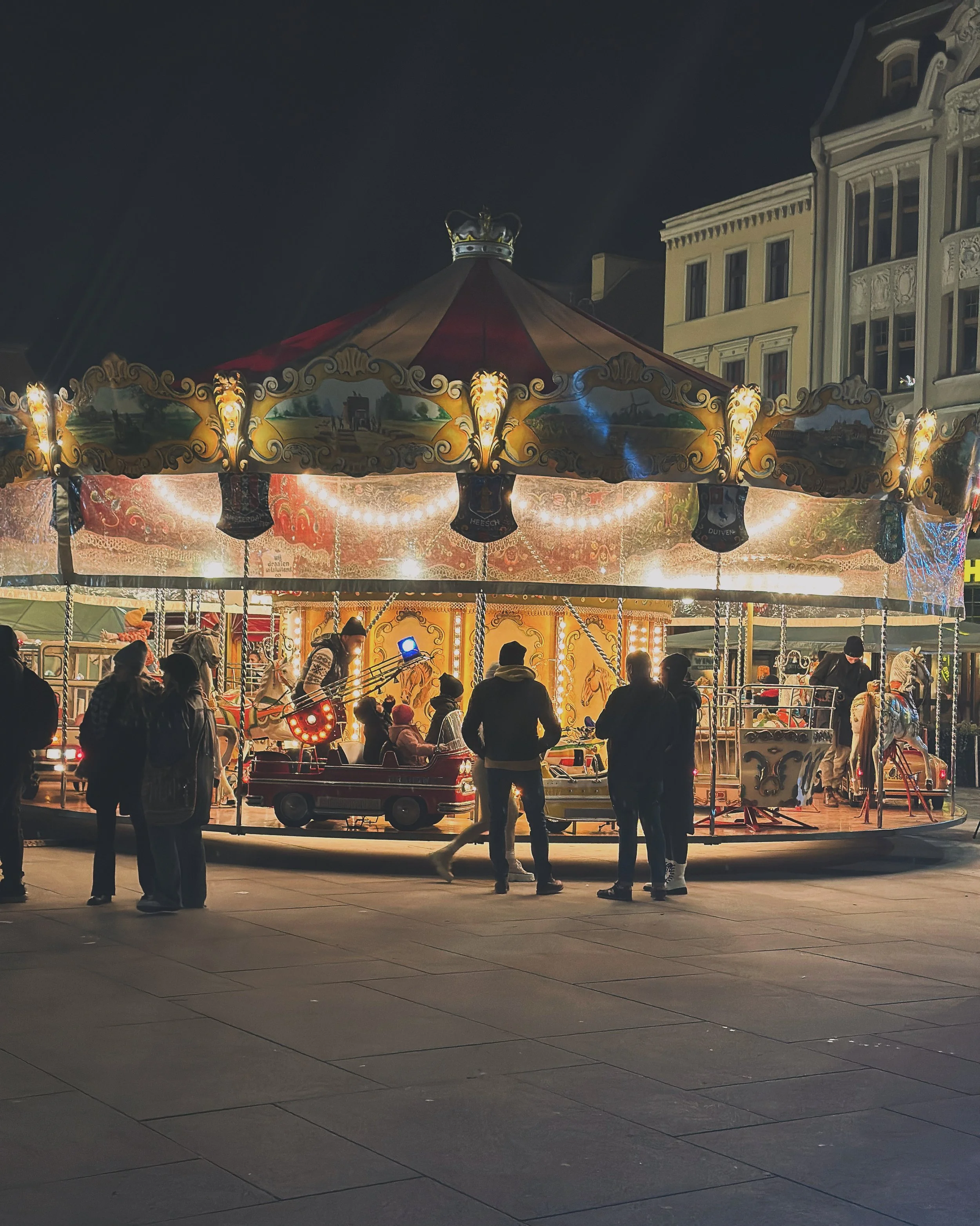 A vintage carousel horse ride at night, illuminated by lights, with people gathered around and buildings in the background.