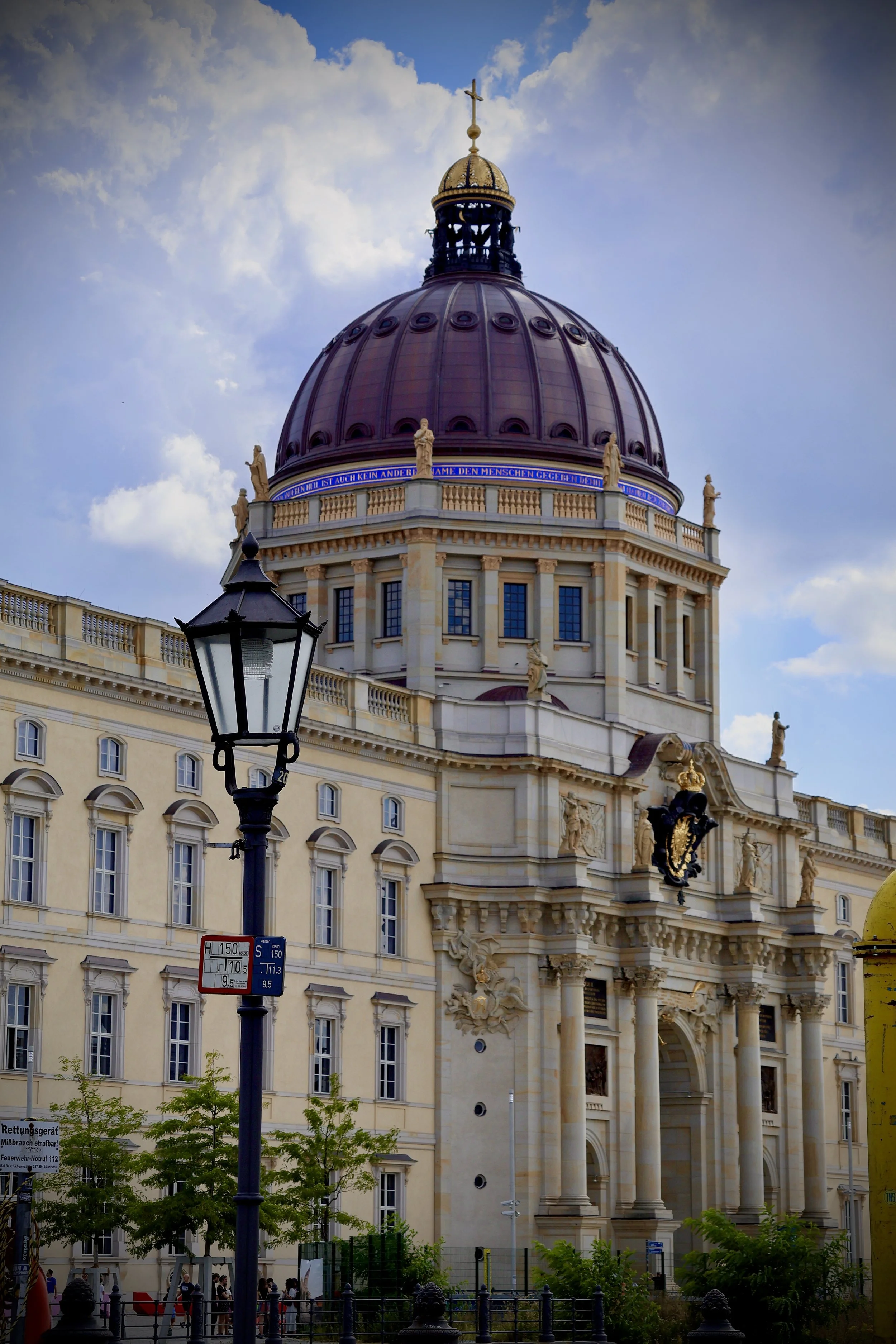 View of a historic European building with a large dome topped by a cross, decorative statues along the roofline, and a street lamp in the foreground. The building has ornate architecture and is set against a partly cloudy sky.