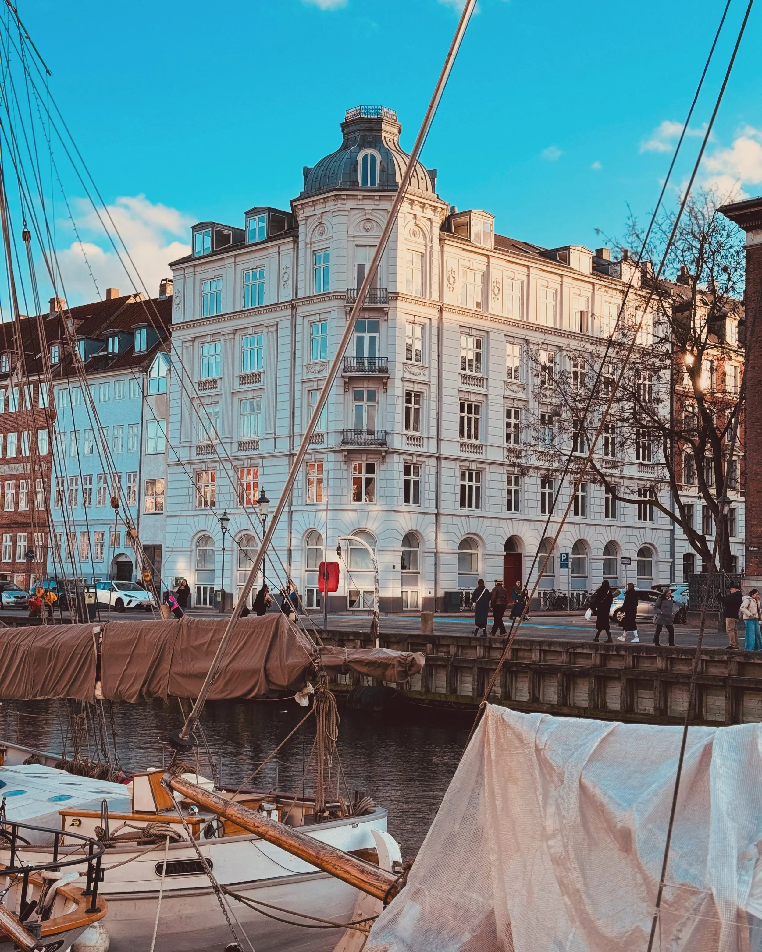 A harbor scene with boats docked along the water, including a sailboat in the foreground covered with a tarp, and a large white multi-story building with ornate architectural details and a rounded tower in the background, against a bright blue sky wi