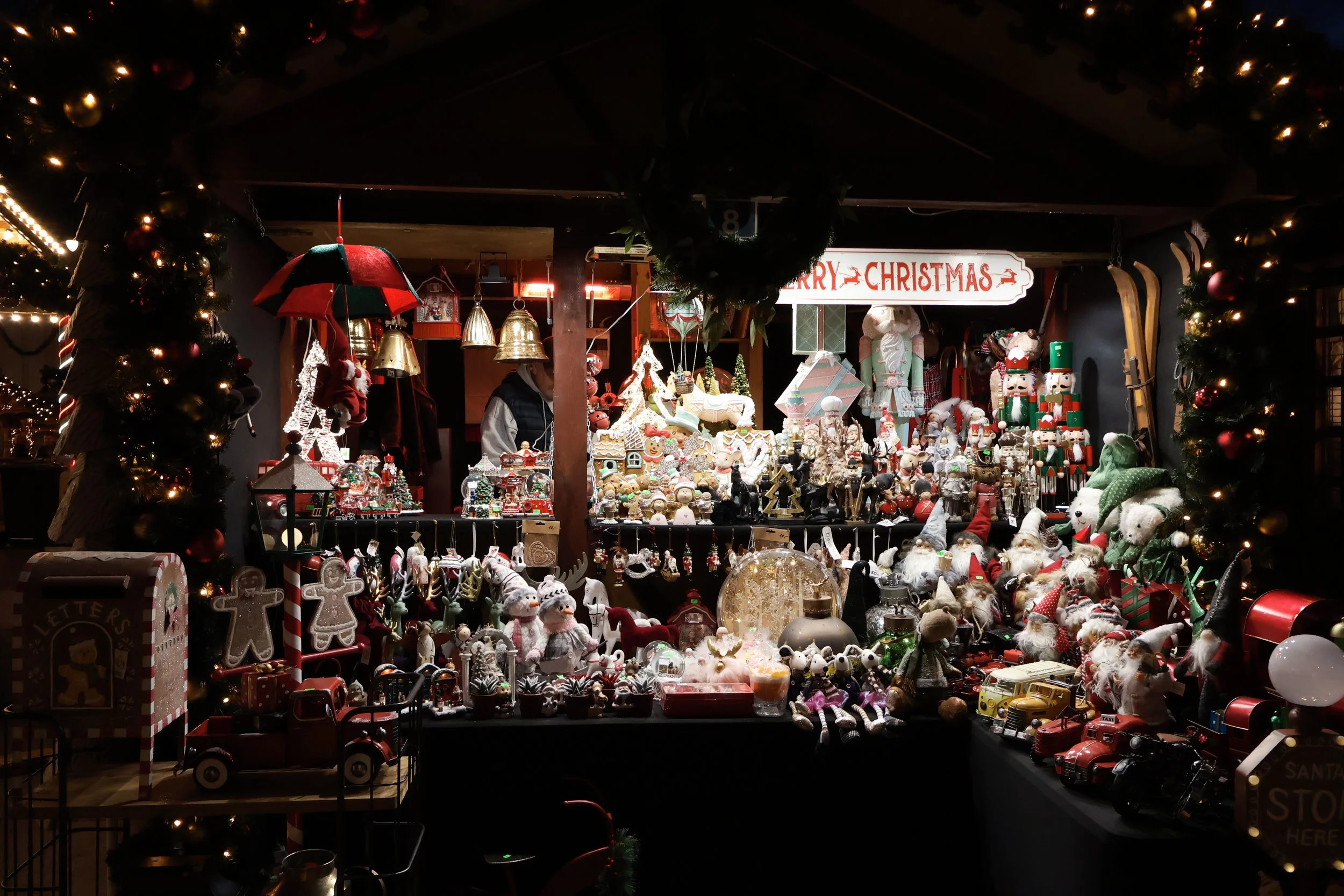 A festive Christmas market stall decorated with Christmas lights, garlands, and ornaments. The stall displays various Christmas decorations and toys, including Santa figurines, gingerbread houses, nutcrackers, and stuffed animals. A large red and whi