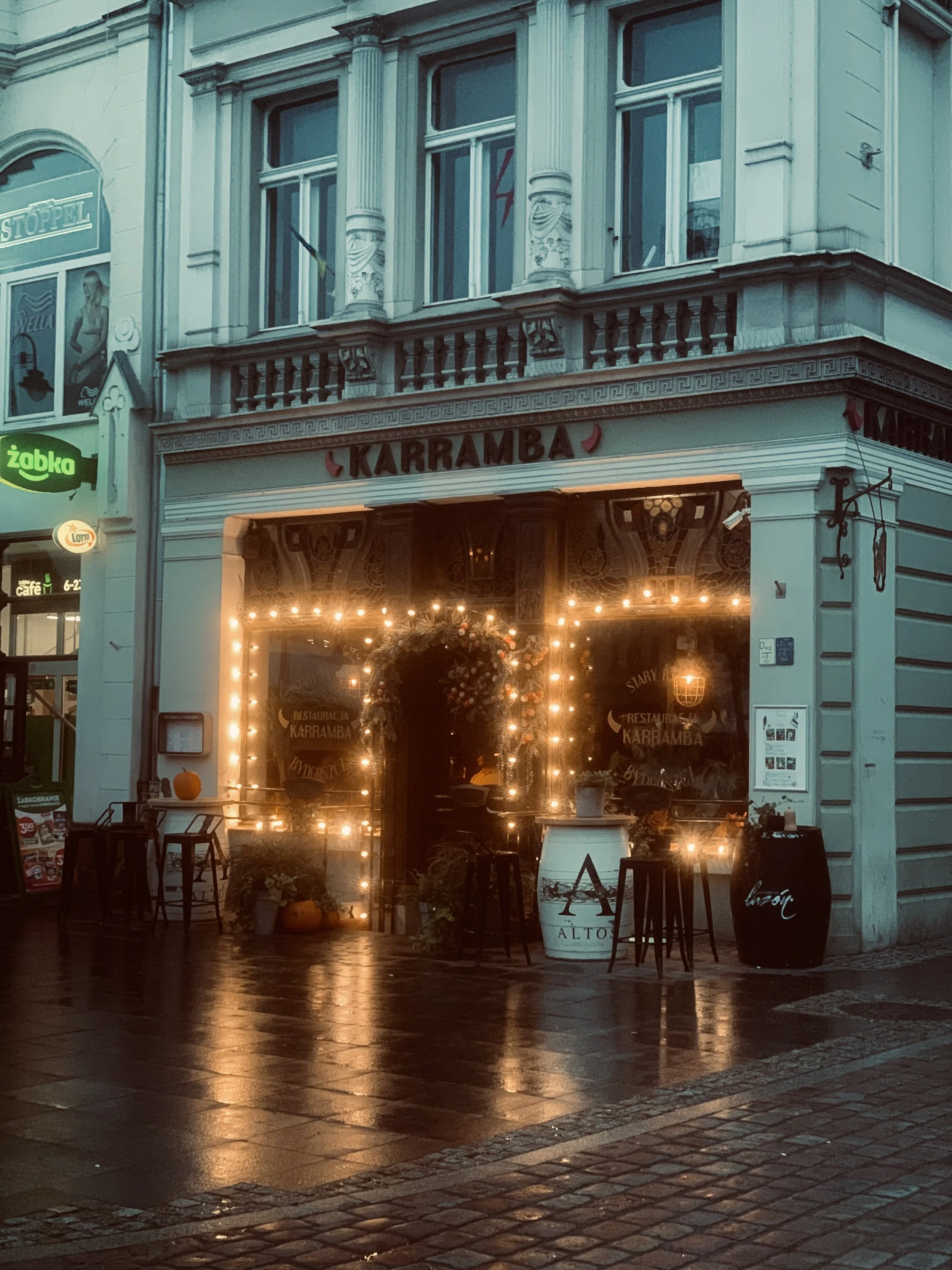 The image shows a building with the sign 'Karramba' above the entrance, decorated with string lights. The storefront has large windows and seating outside, and the street appears wet, reflecting the lights.