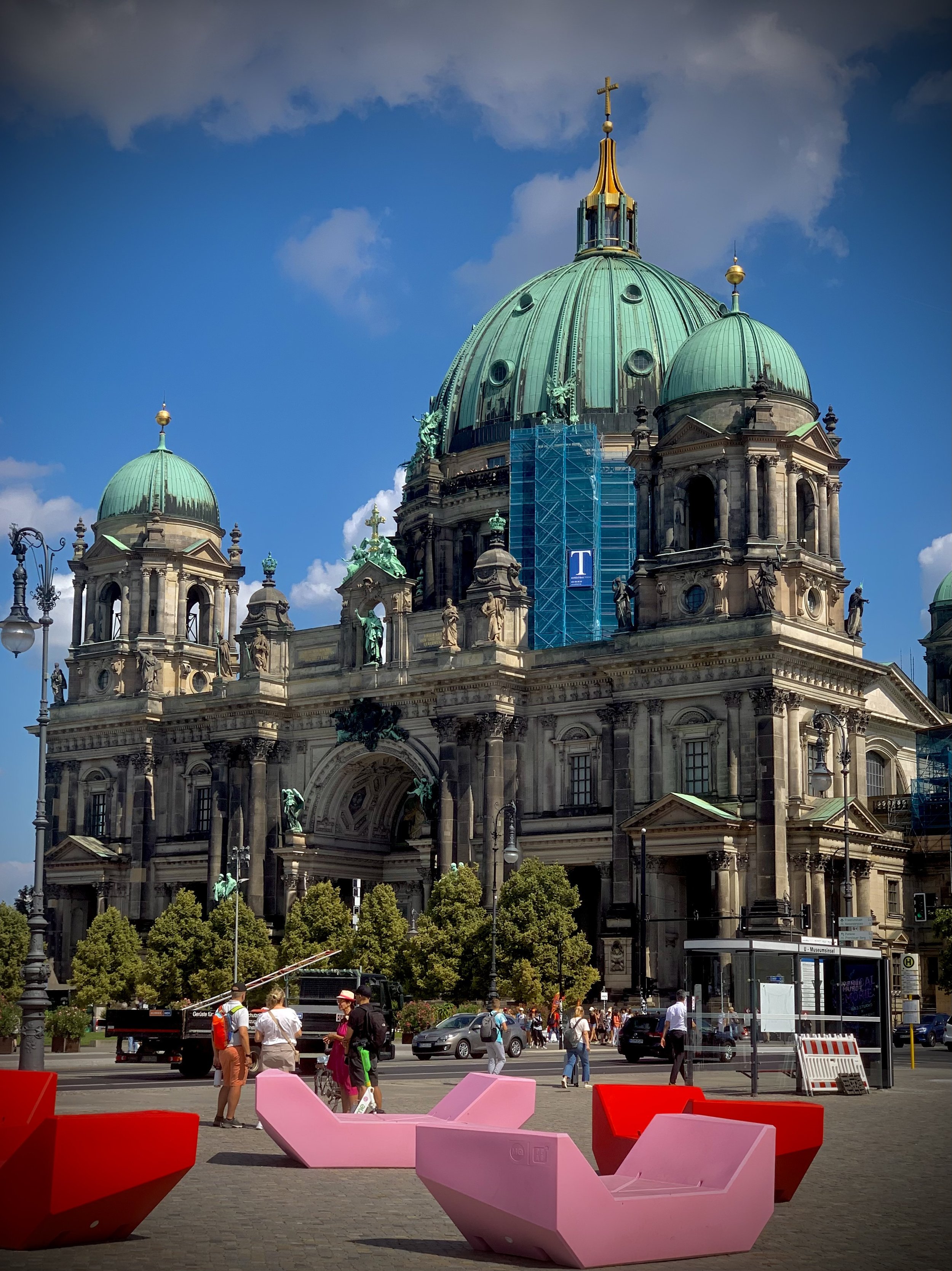 A historic building with a large green dome, smaller green domes, and ornate architectural details, with trees and a busy city street with people and cars in the foreground.