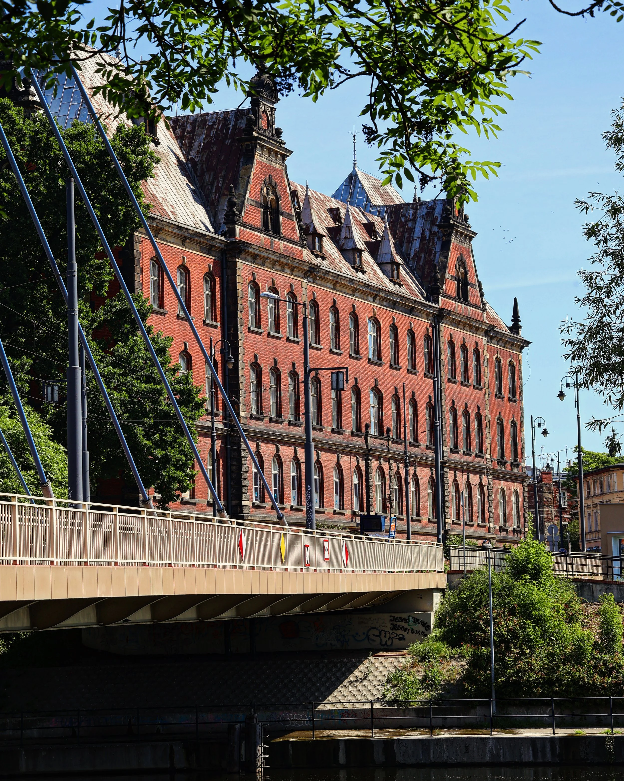 View of a historic red brick building with intricate architectural details, multiple gabled roofs with rusted metal, surrounded by green trees and street lamps, beneath a bright blue sky.