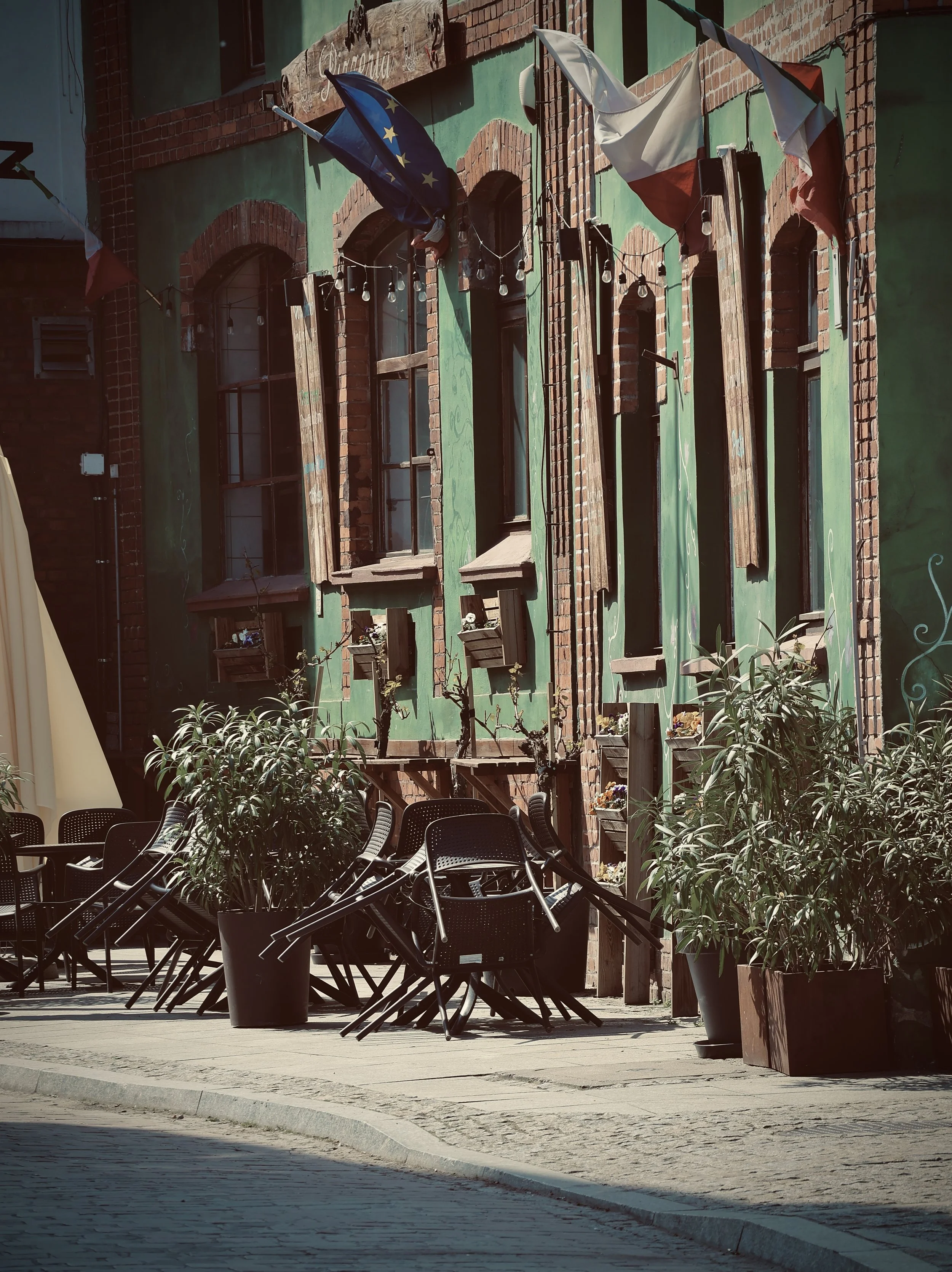 Empty outdoor patio with black chairs stacked, potted plants, and a brick and green painted building with flags and string lights hanging.