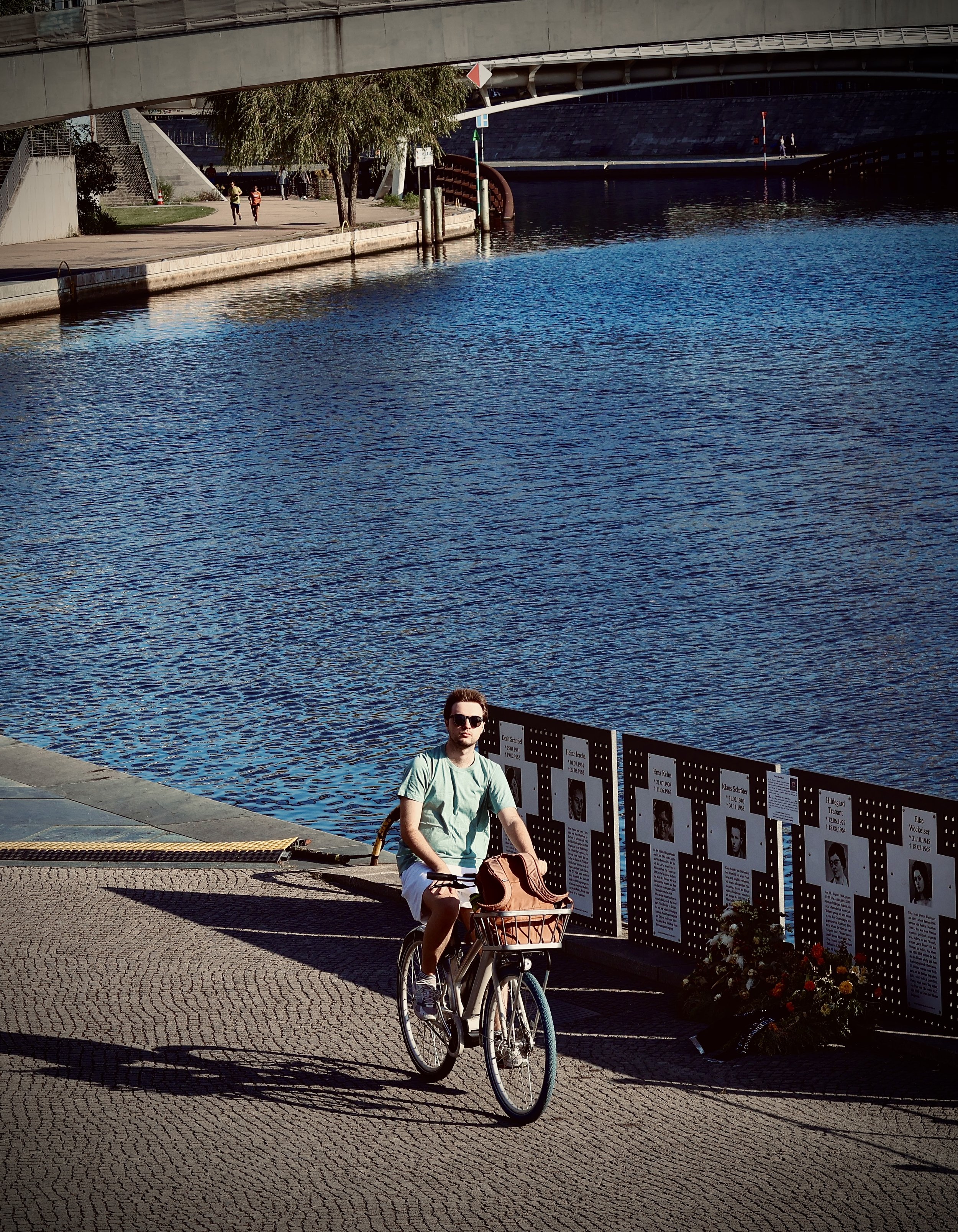 A young man wearing sunglasses and a light green T-shirt sitting on a white bicycle with a basket over the front while posing beside a memorial wall of photographs and names along a historic waterfront promenade in the late afternoon.