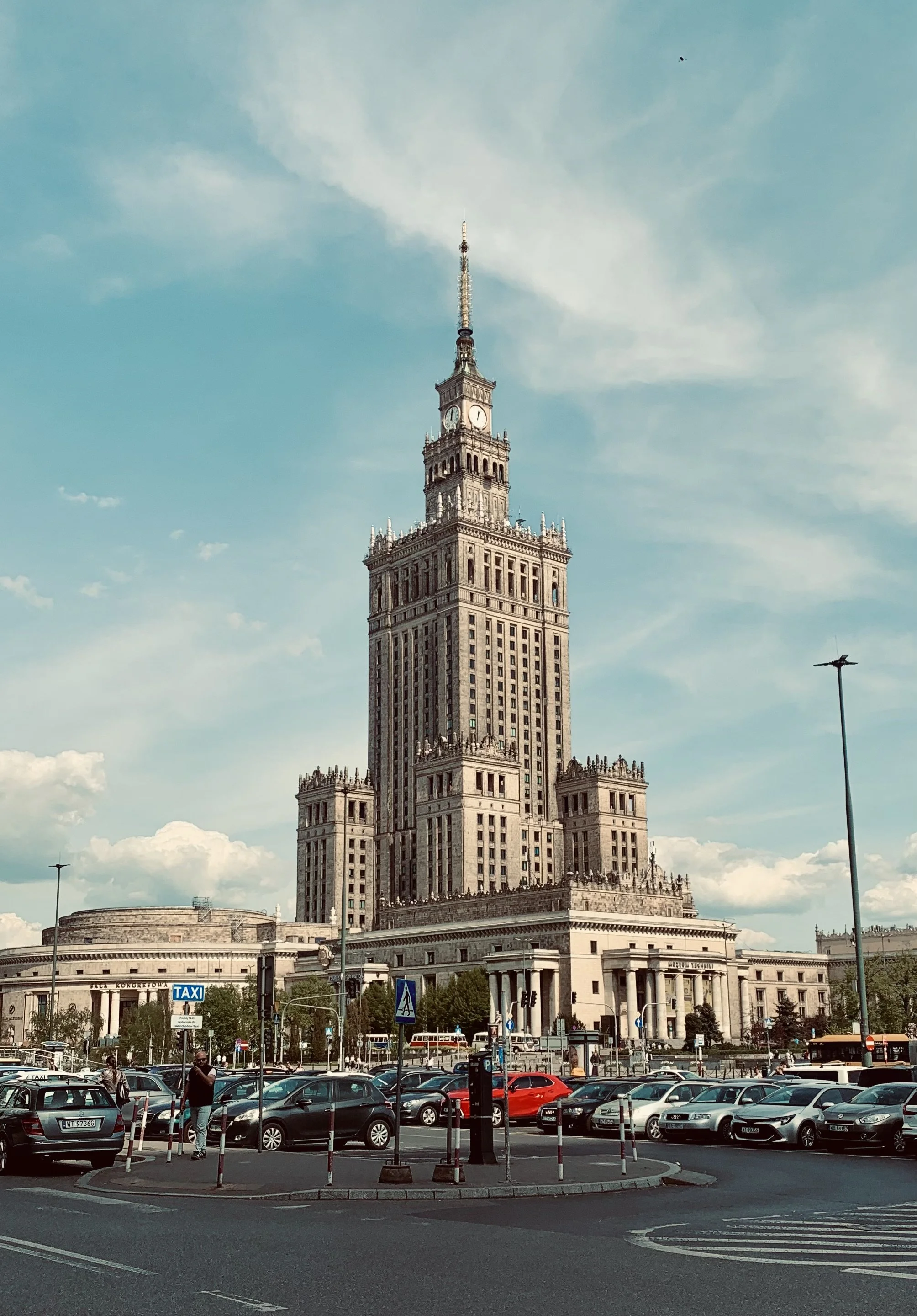 The Palace of Culture and Science, a tall historic skyscraper with a clock on the top, situated in a busy city area with cars and pedestrians, under a partly cloudy sky.