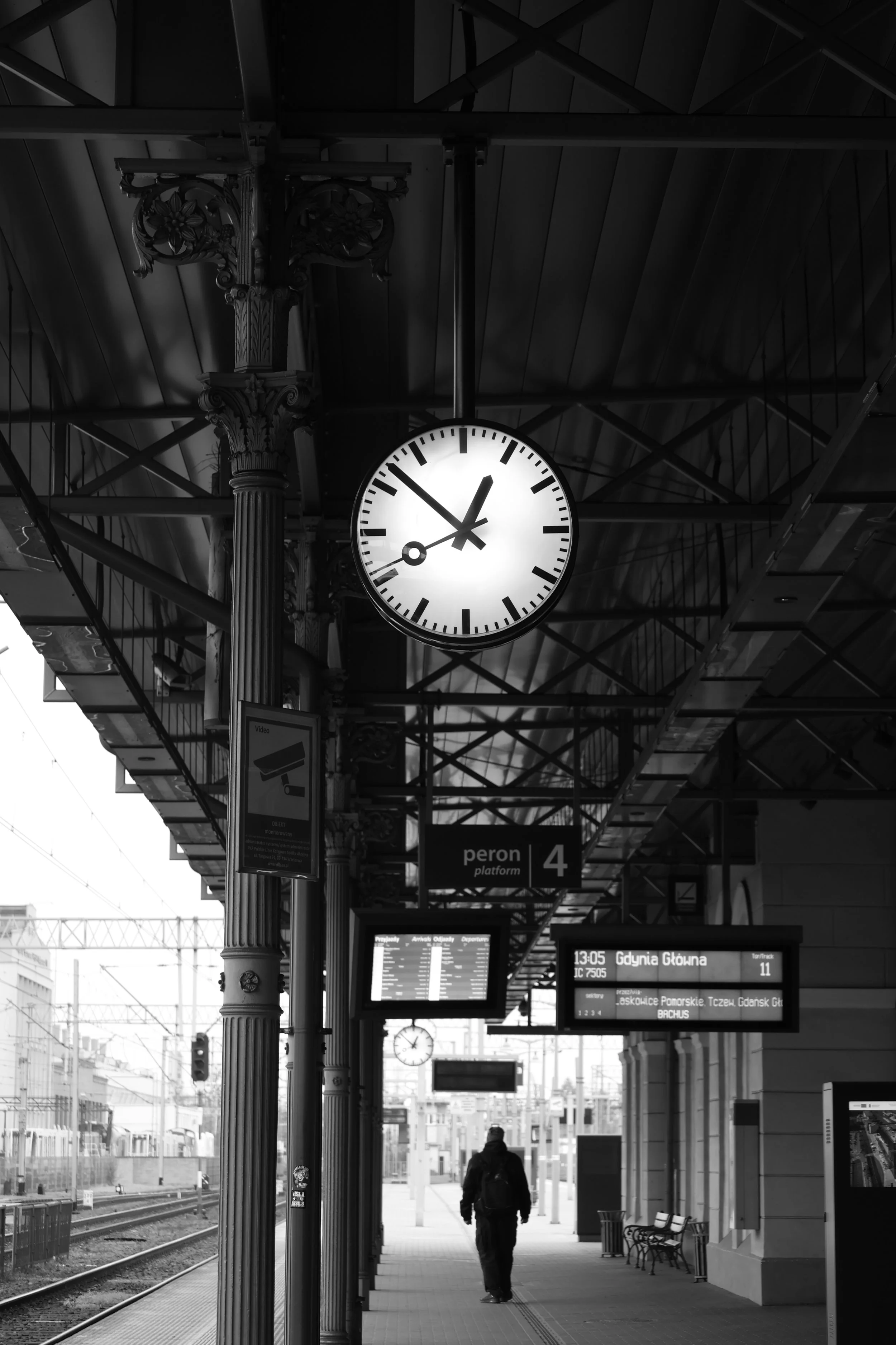 A black and white photo of a train station platform with a decorative column supporting a clock showing the time as 1:50. Digital and electronic display boards are visible with train information and schedules, and there is a solitary person walking a