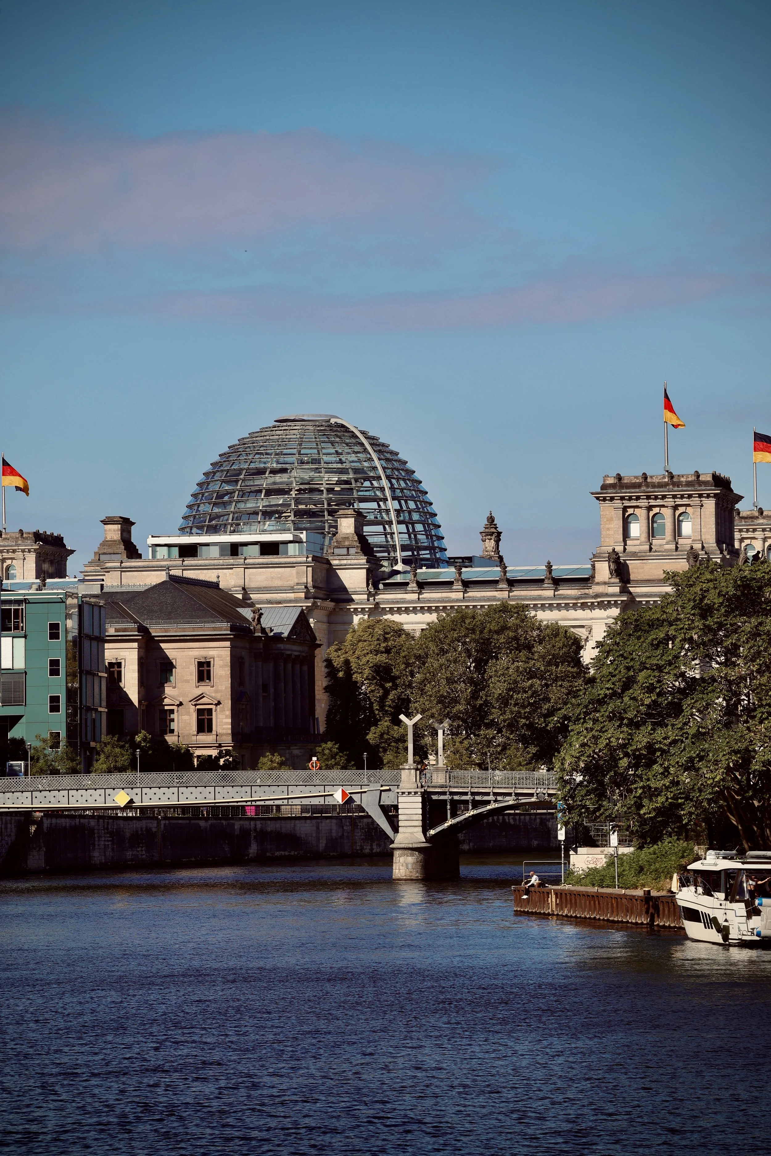 View of Berlin skyline with the Reichstag building and the glass dome, flags on the rooftops, river in the foreground, and trees along the riverbank on a clear day.