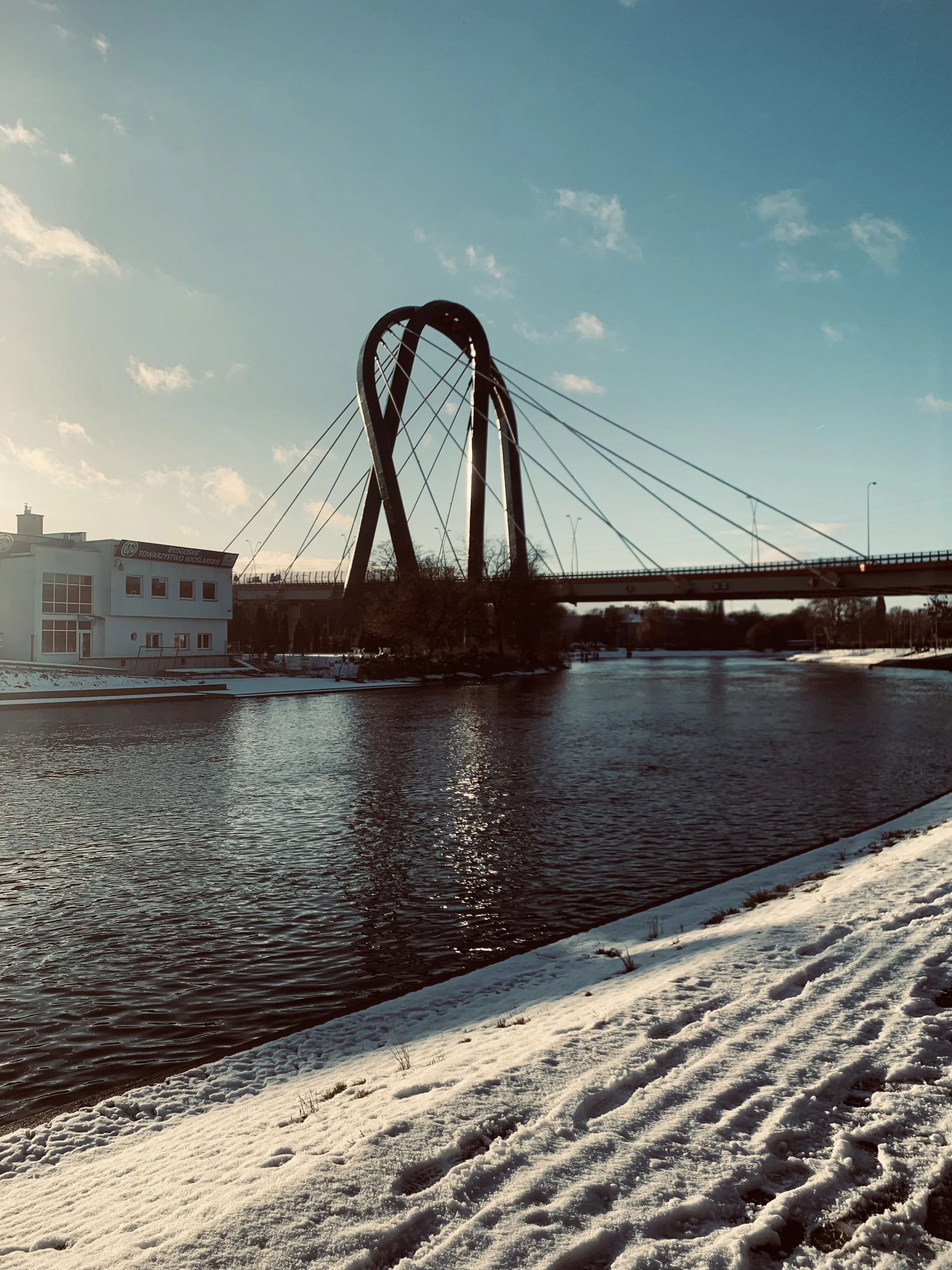 A river with snow along its banks, a modern arched bridge spans across, with a building and trees nearby under a clear blue sky.