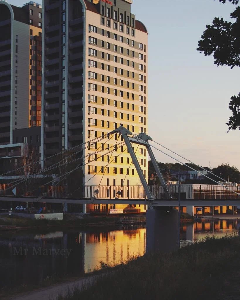 High-rise building next to a waterway with a modern bridge and trees in the foreground during sunset.
