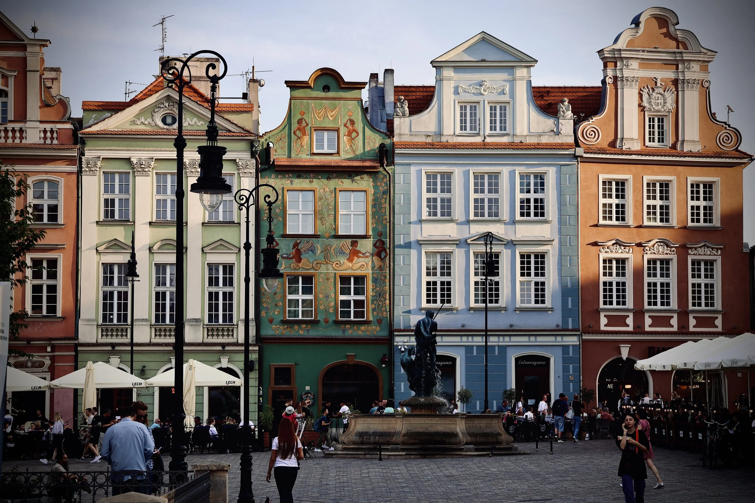 Colorful historic buildings with decorative facades lining a busy outdoor plaza with people dining and walking, a central fountain, lampposts, and umbrellas.