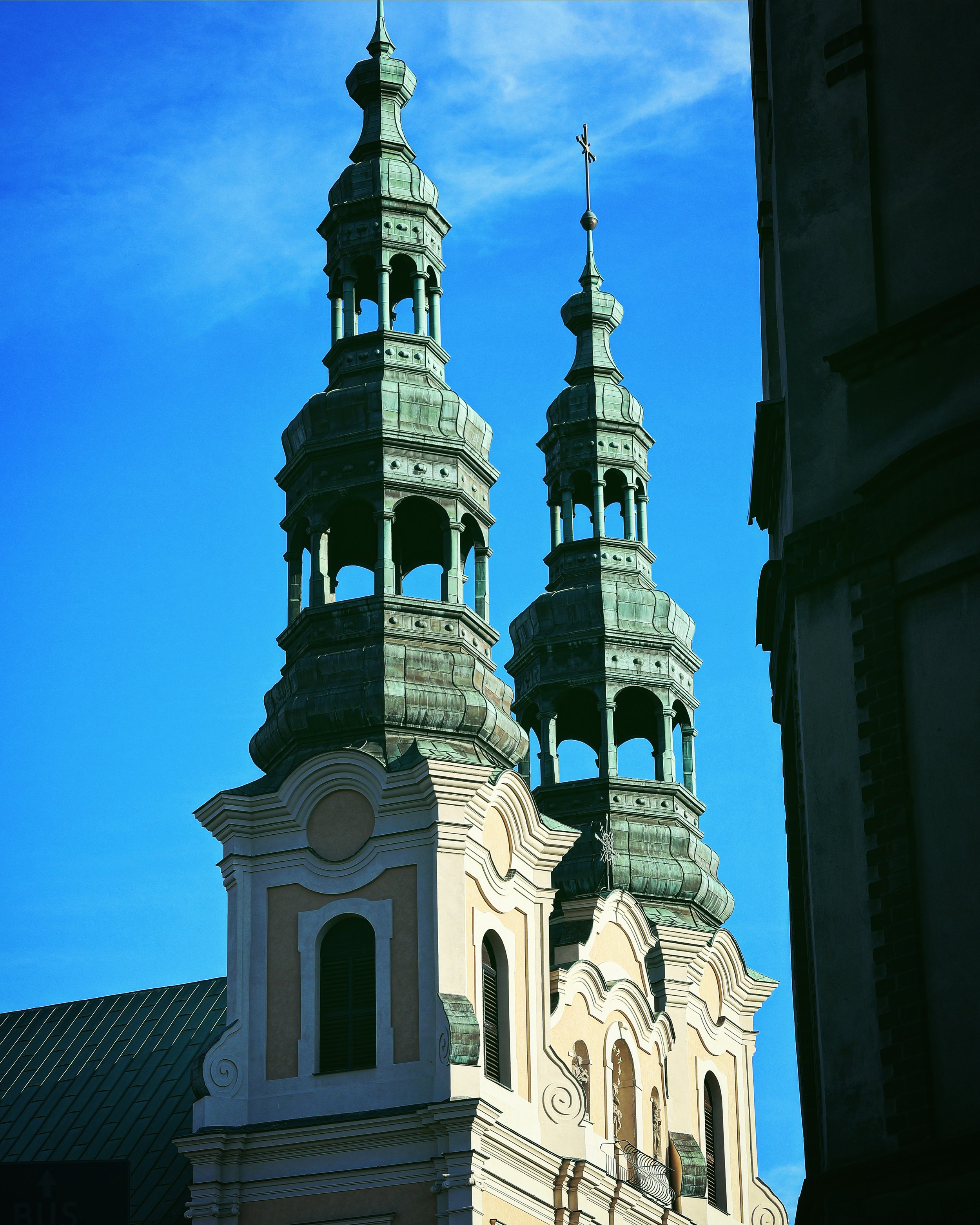 The image depicts the upper part of a Baroque or Rococo style church with ornate spires and a cross on top, against a bright blue sky. The church has detailed architectural elements, including curved pediments, column-like structures, and decorative 