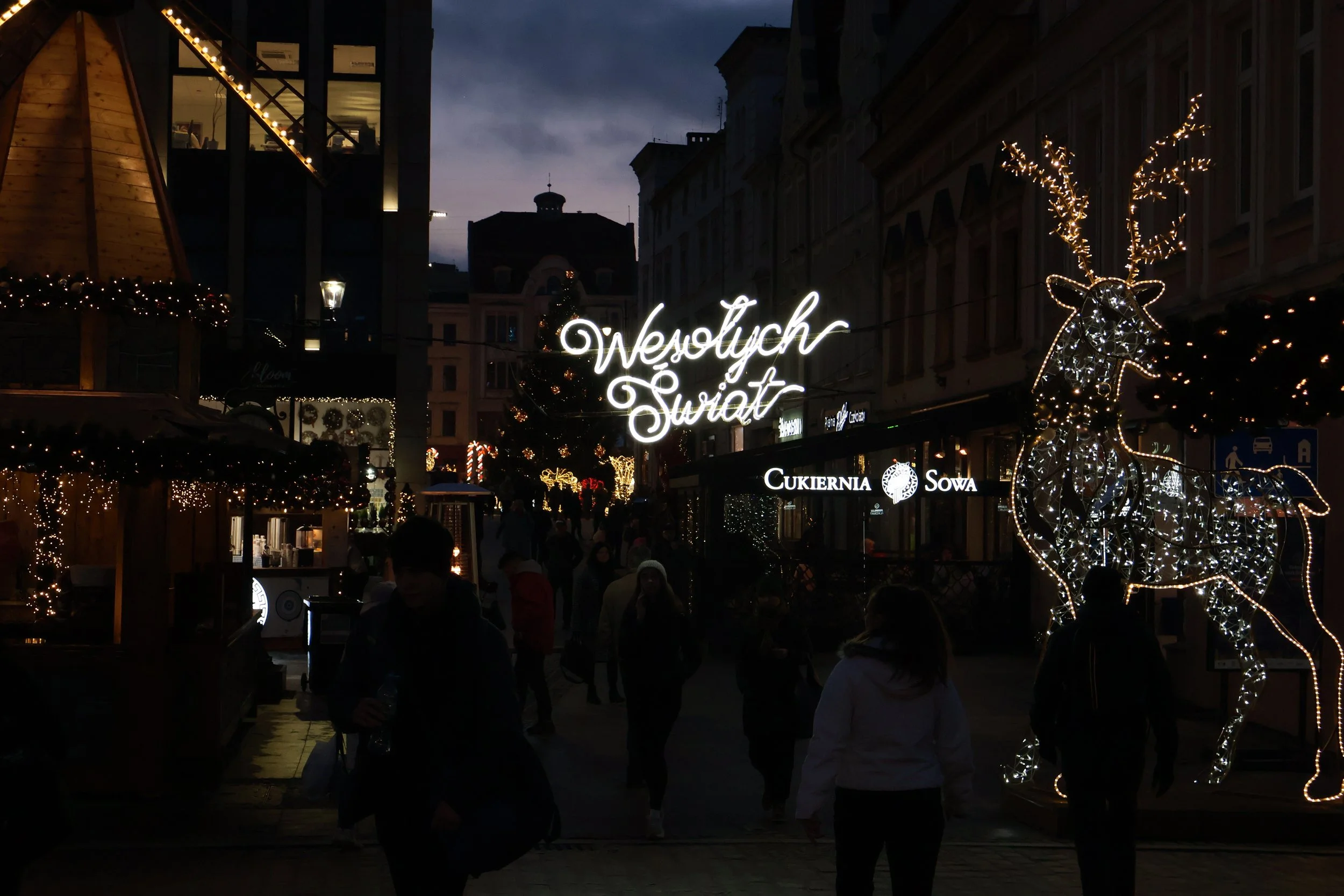 Nighttime Christmas market scene with festive lights, including a glowing reindeer figure, illuminated sign saying 'Wesołych Świąt,' and decorated buildings.