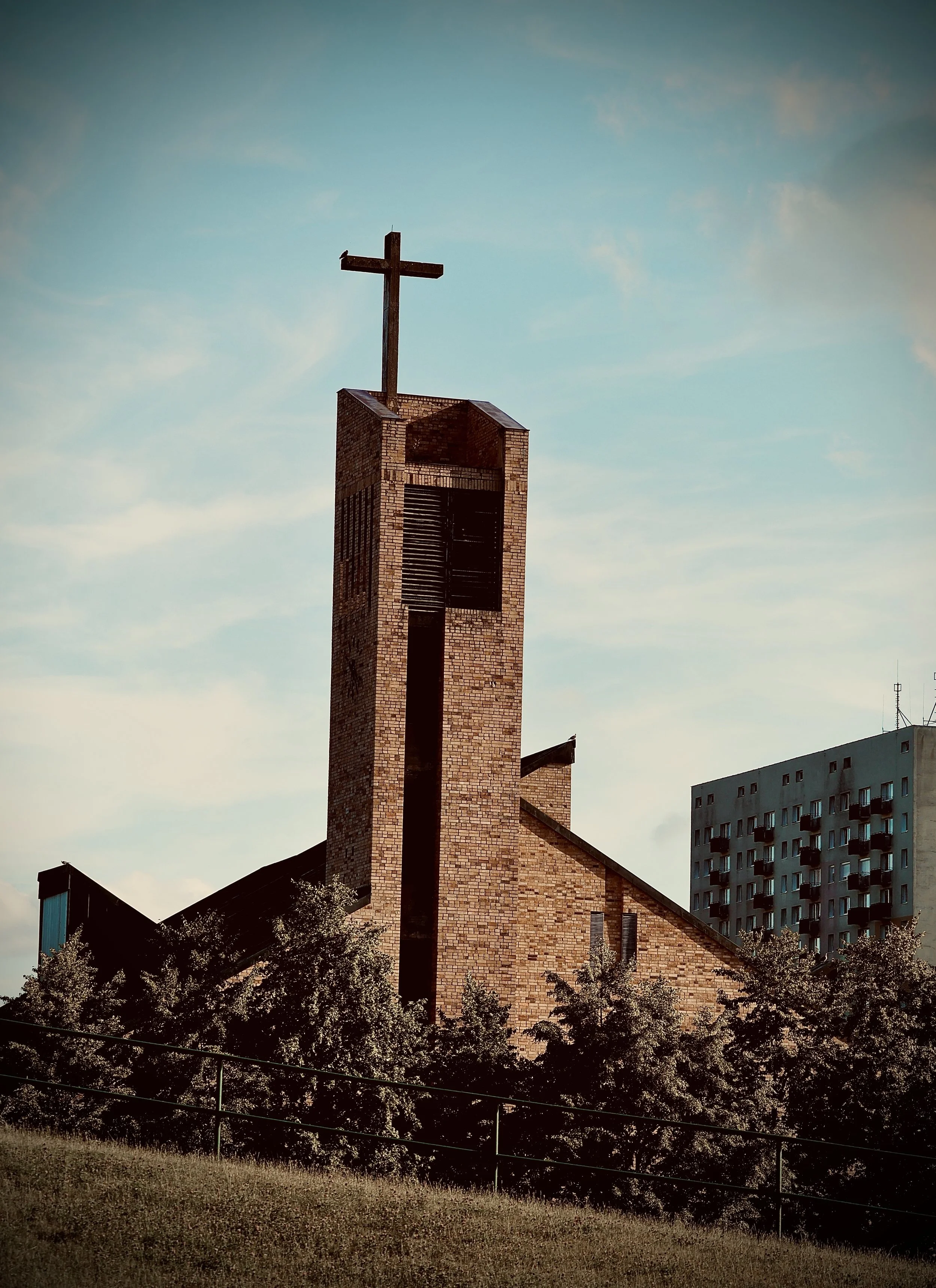 A brick church with a tall steeple and a cross on top, surrounded by trees and a fence, with a modern apartment building in the background, under a blue sky with some clouds.