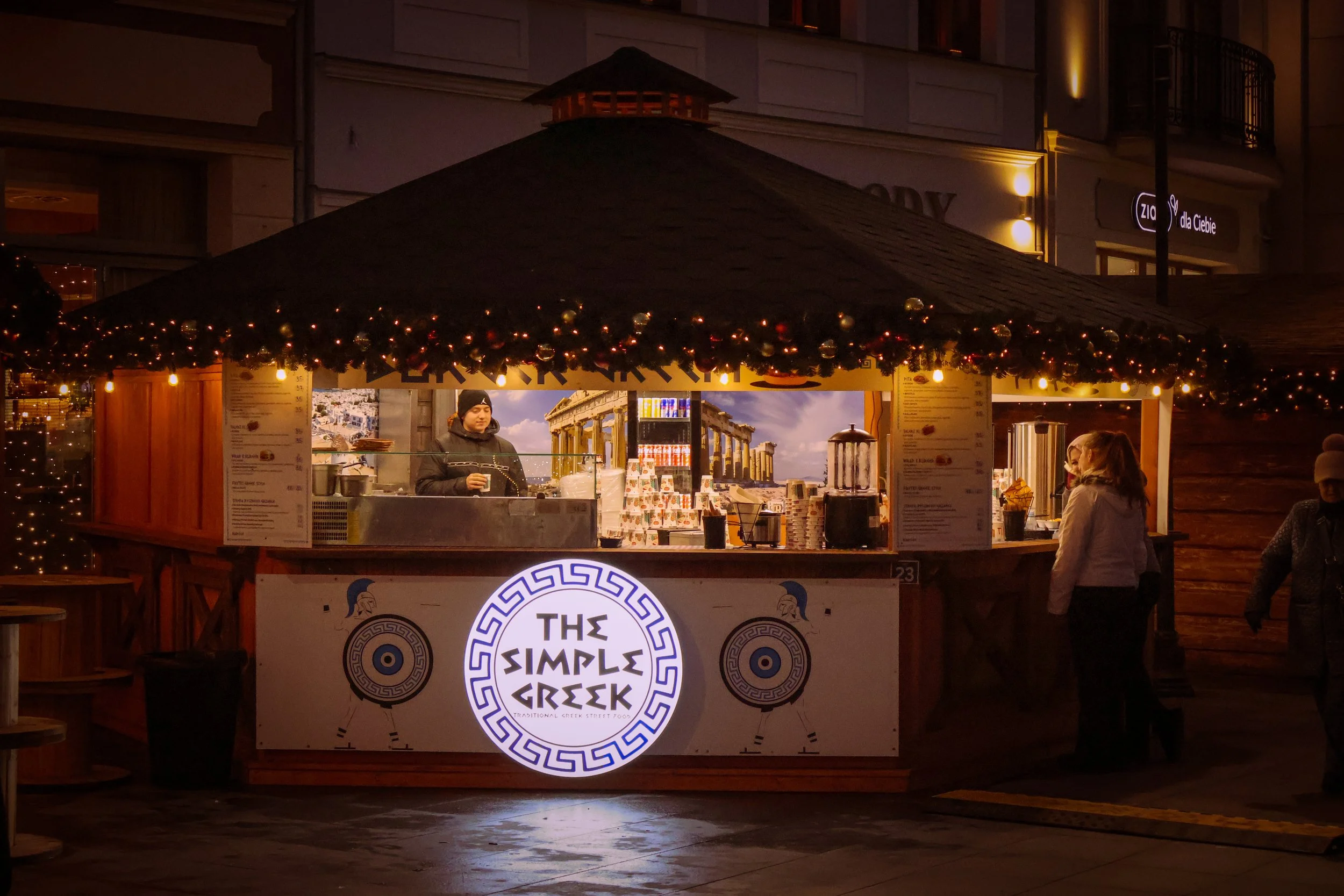 A nighttime scene at an outdoor Greek street food stall decorated with Christmas ornaments. The stall has a sign that reads 'The Simple Greek' and features a Greek-inspired design. There are people standing nearby, and a person behind the counter app