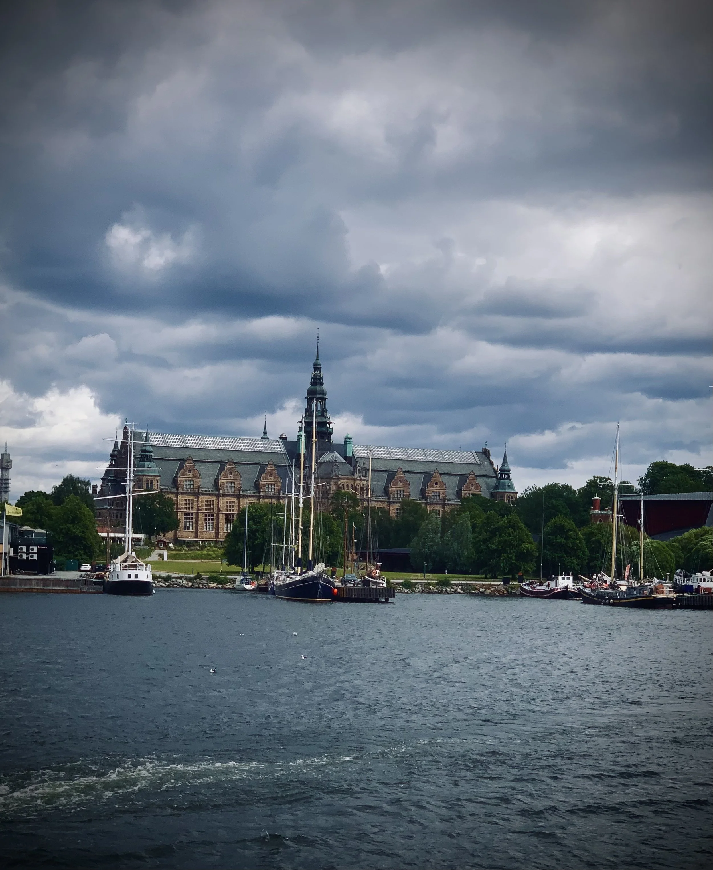 A harbor with sailboats docked and a historic building with a tall spire in the background, under a cloudy sky.